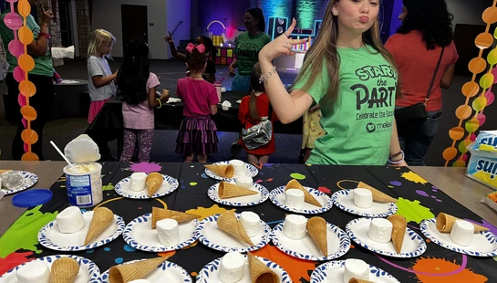 A girl in a green t-shirt poses playfully behind a table set with paper plates holding marshmallows and sugar cones for a party activity.