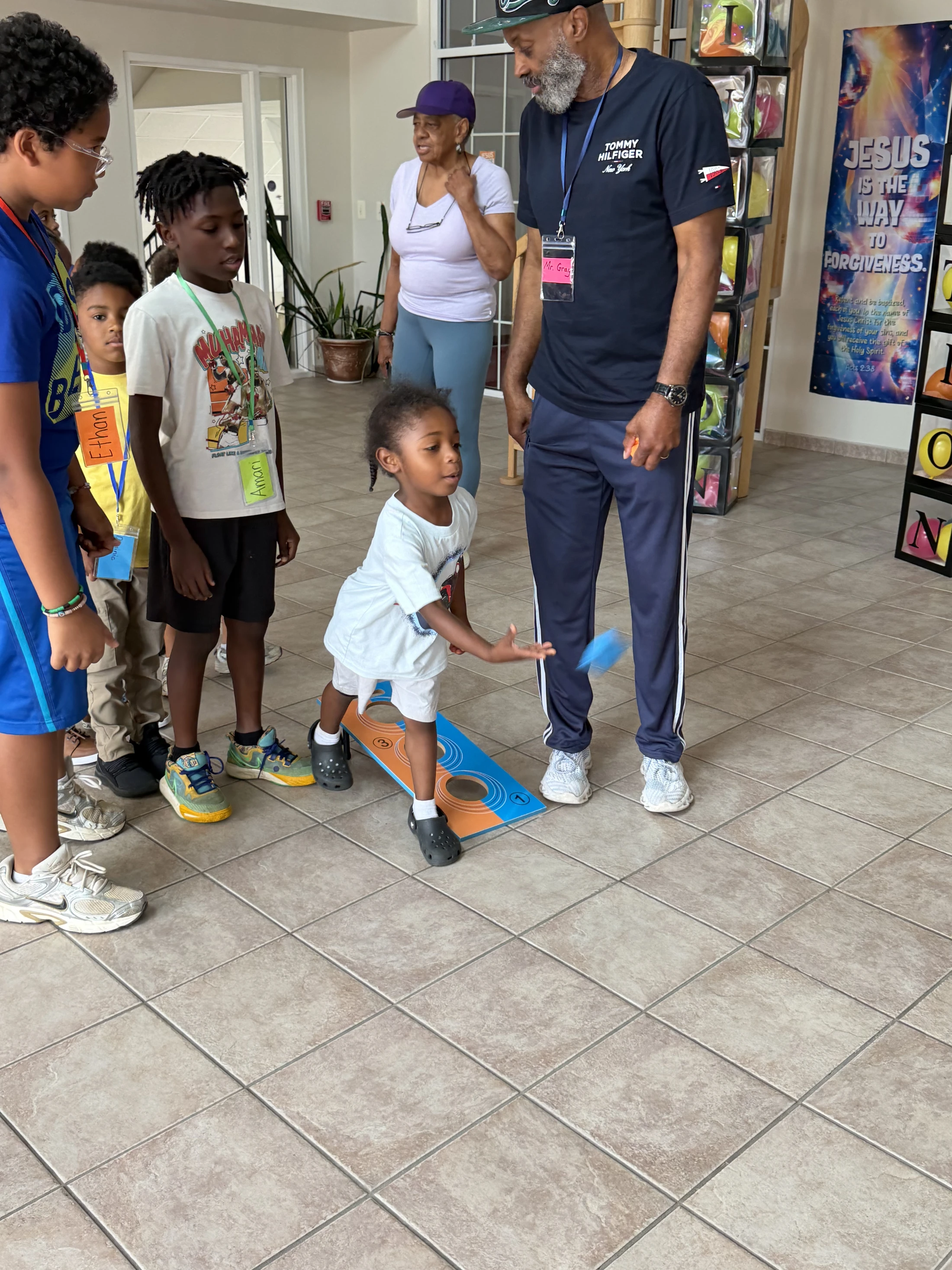 A young Black child throws a bean bag at a cornhole board as other children and two adults watch. They are indoors in a room with a tiled floor.