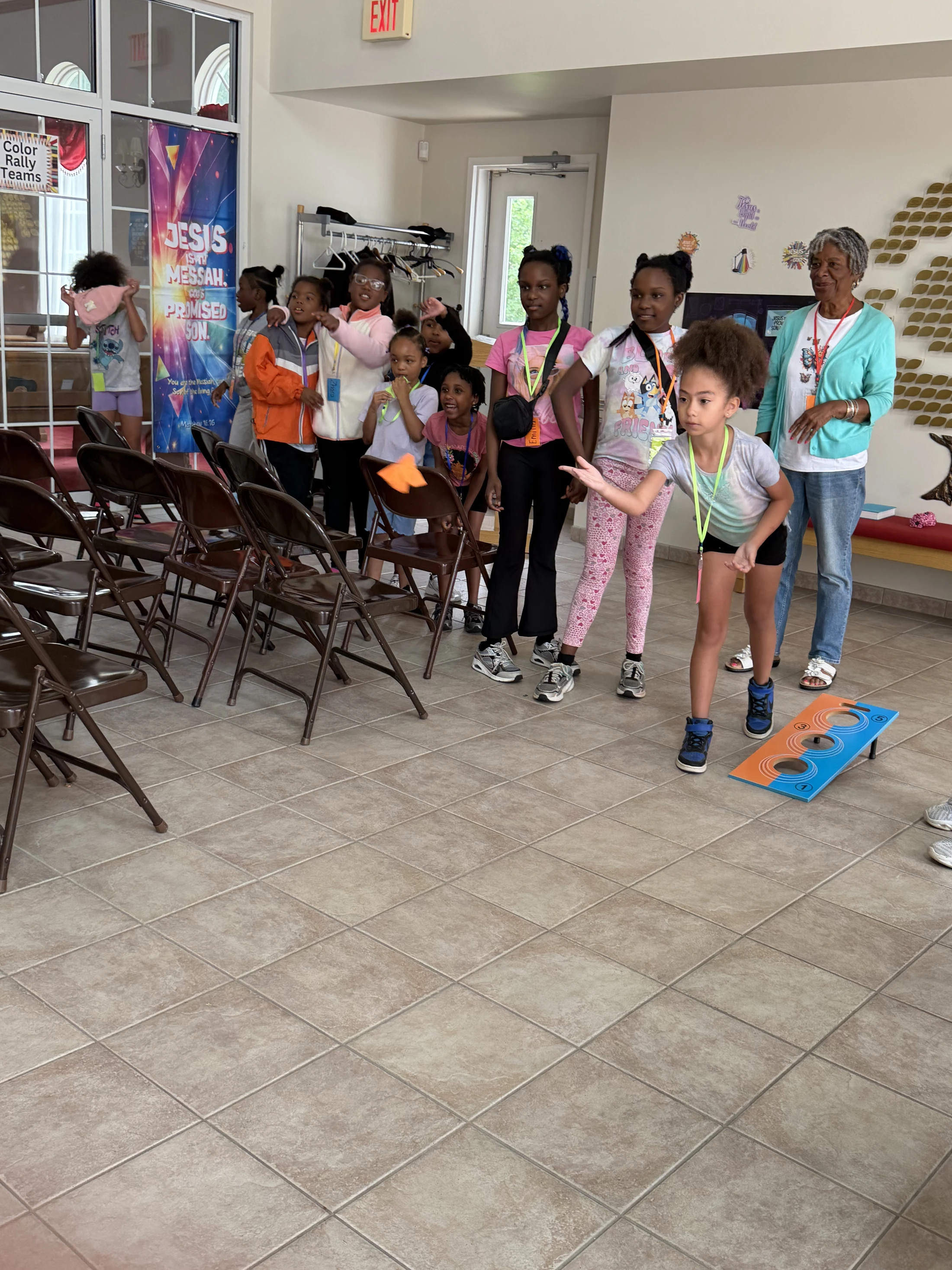A young girl prepares to throw a beanbag at a target game on a tiled floor, as a group of other children and an adult watch in a community room.