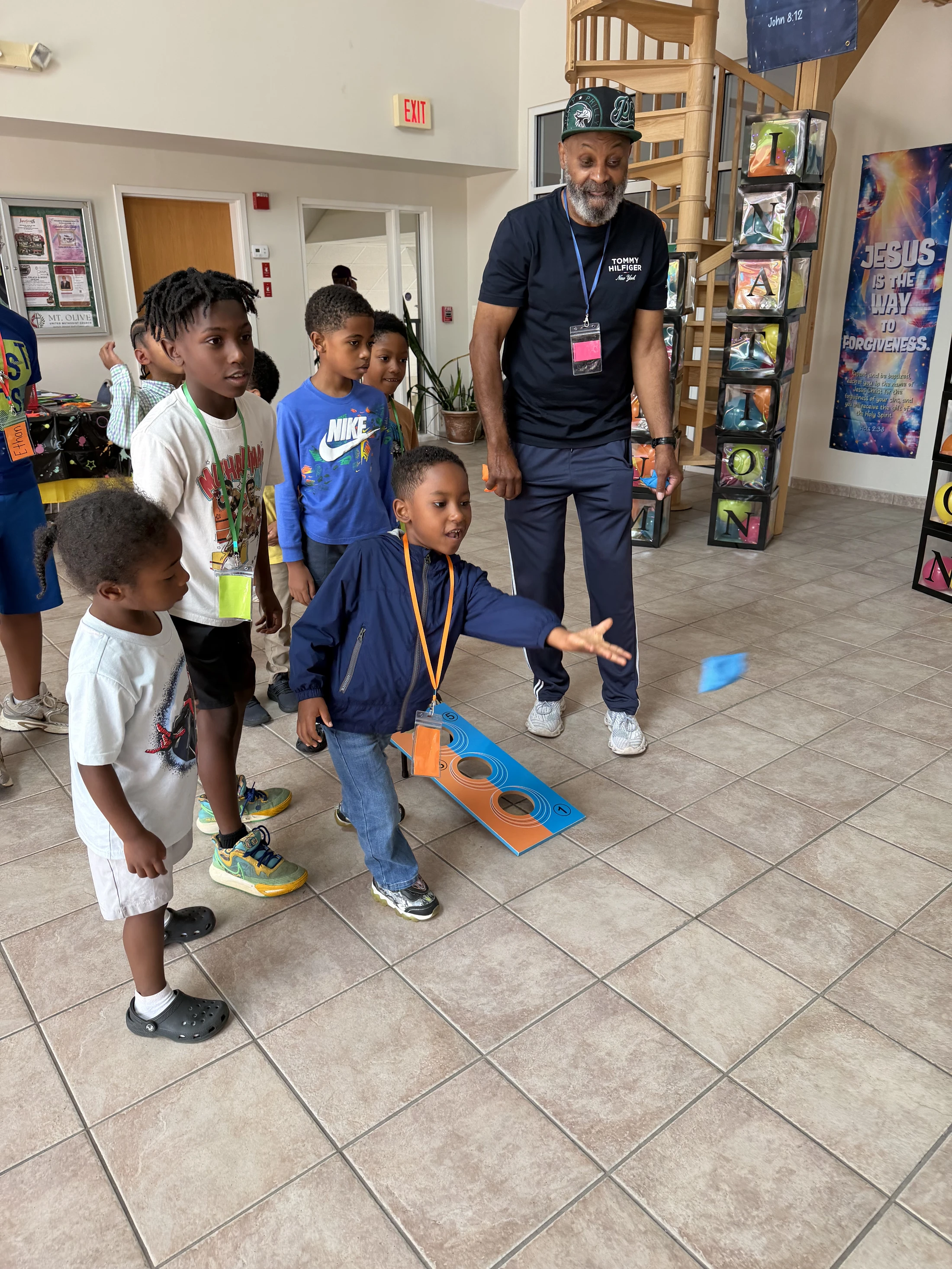 An older Black man smiles as he watches a group of young Black children play a beanbag toss game indoors. A boy in a blue jacket is mid-throw.