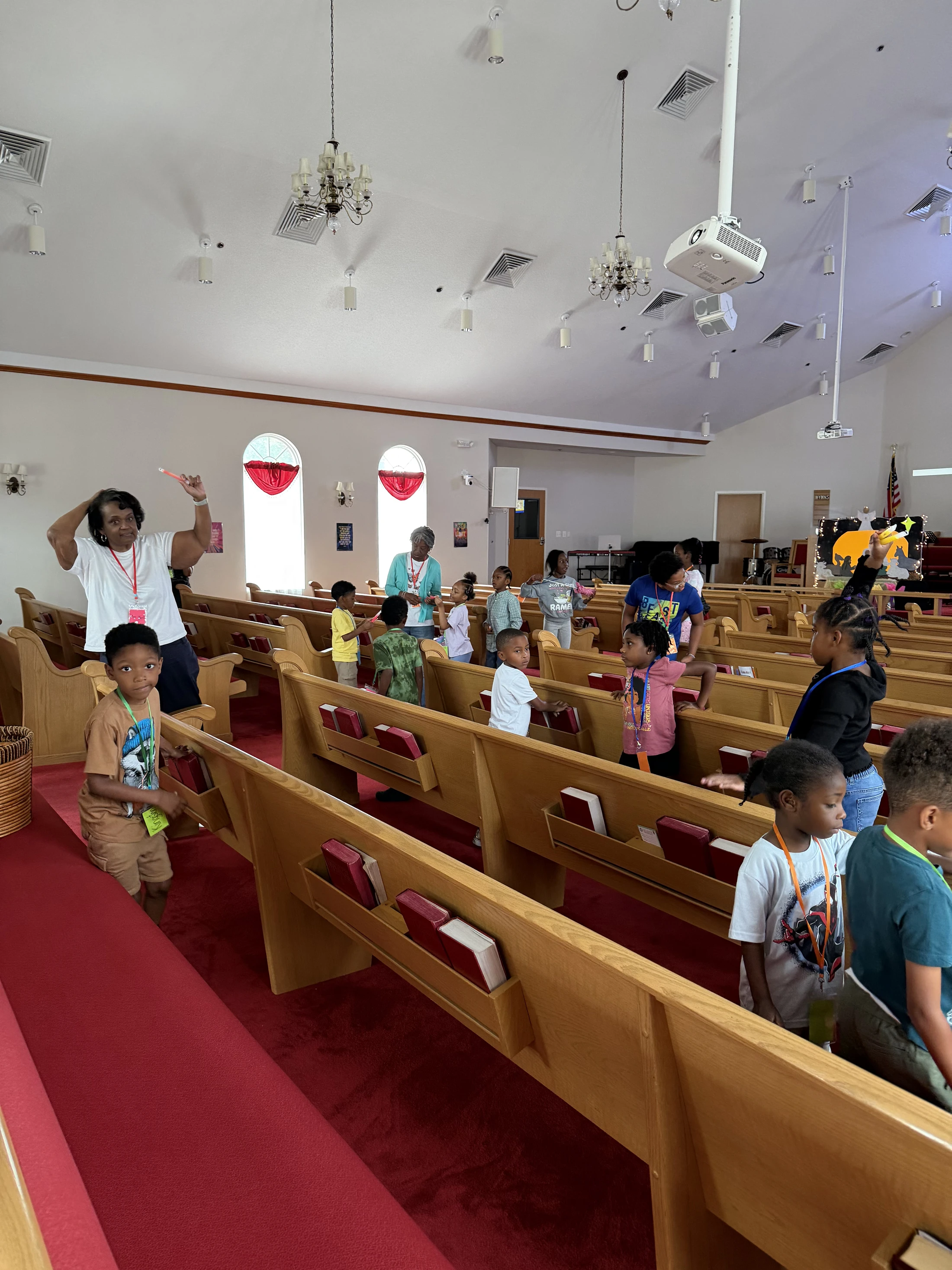 A group of children and a few adults stand between the wooden pews in a church sanctuary. A woman in a white shirt gestures as if leading an activity.