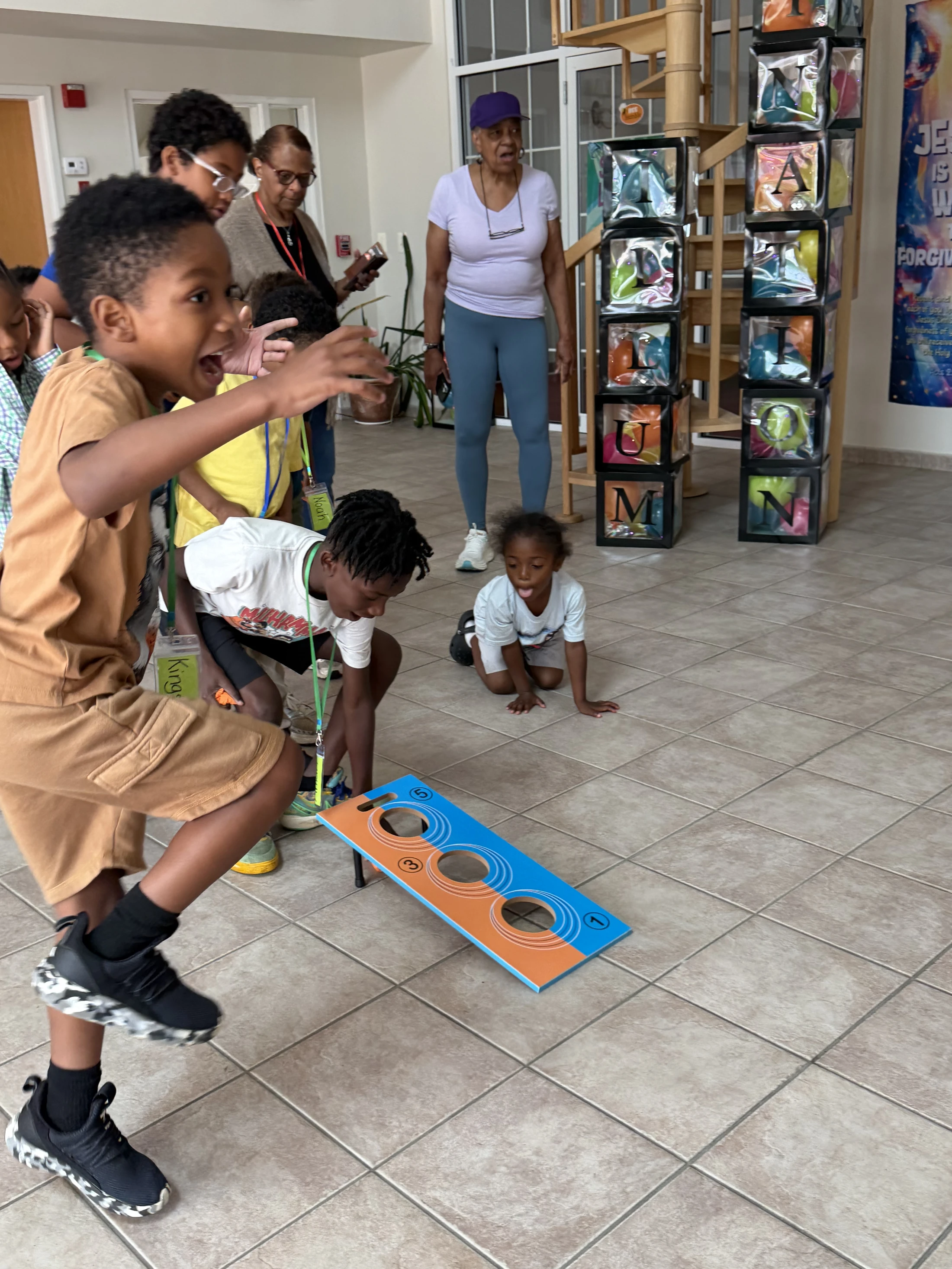 An excited young boy in a brown outfit jumps while playing an indoor toss game. Other children and adults watch as a toddler crawls on the tiled floor nearby.