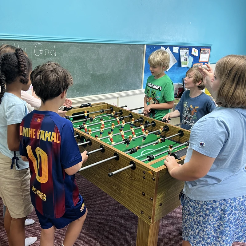 A group of children stand around a wooden foosball table in a classroom with blue walls, actively engaged in playing a game together.