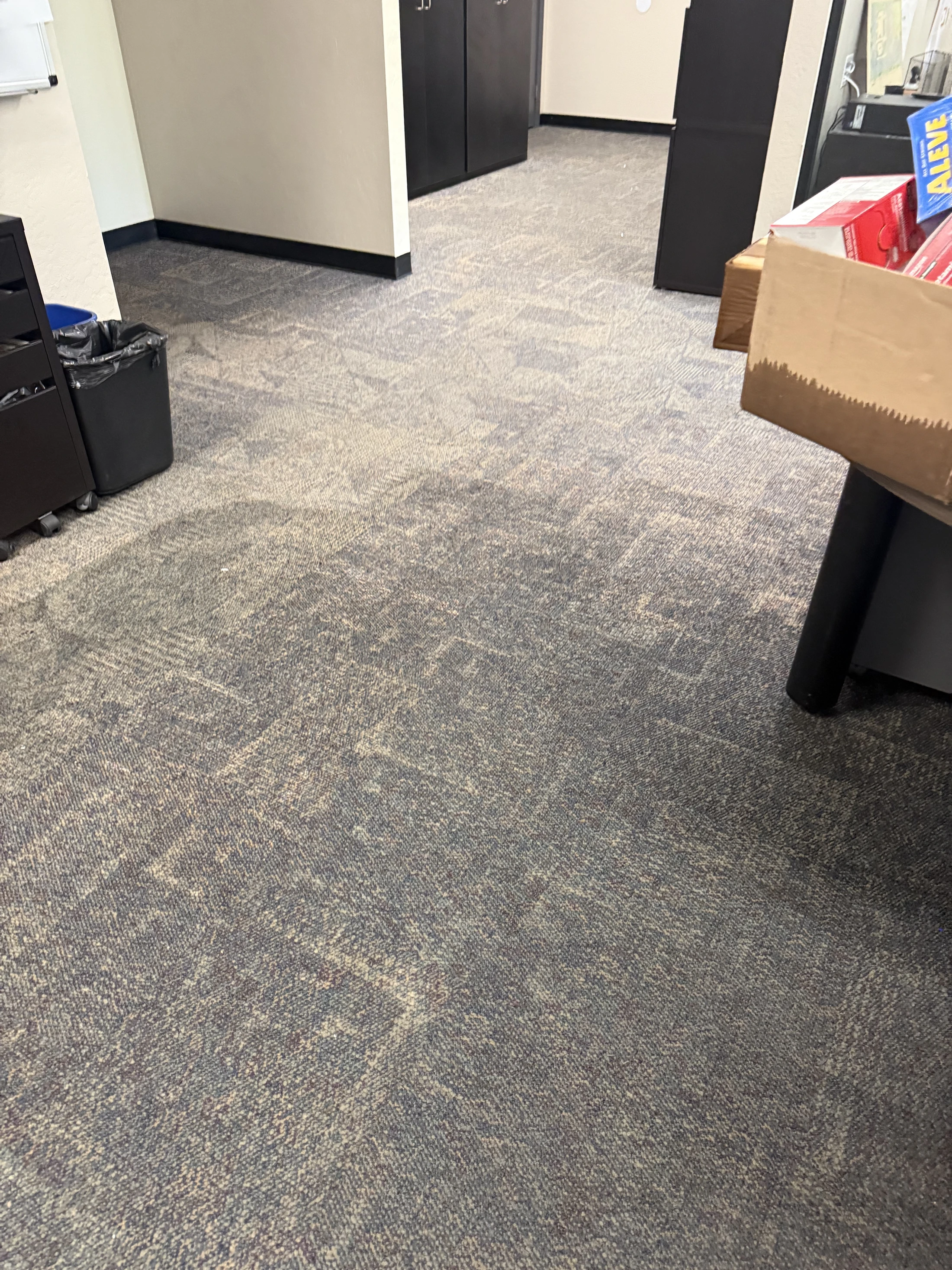 A view of a gray patterned carpet in an office setting, with a black trash can, cabinets, and a desk corner visible in the background.