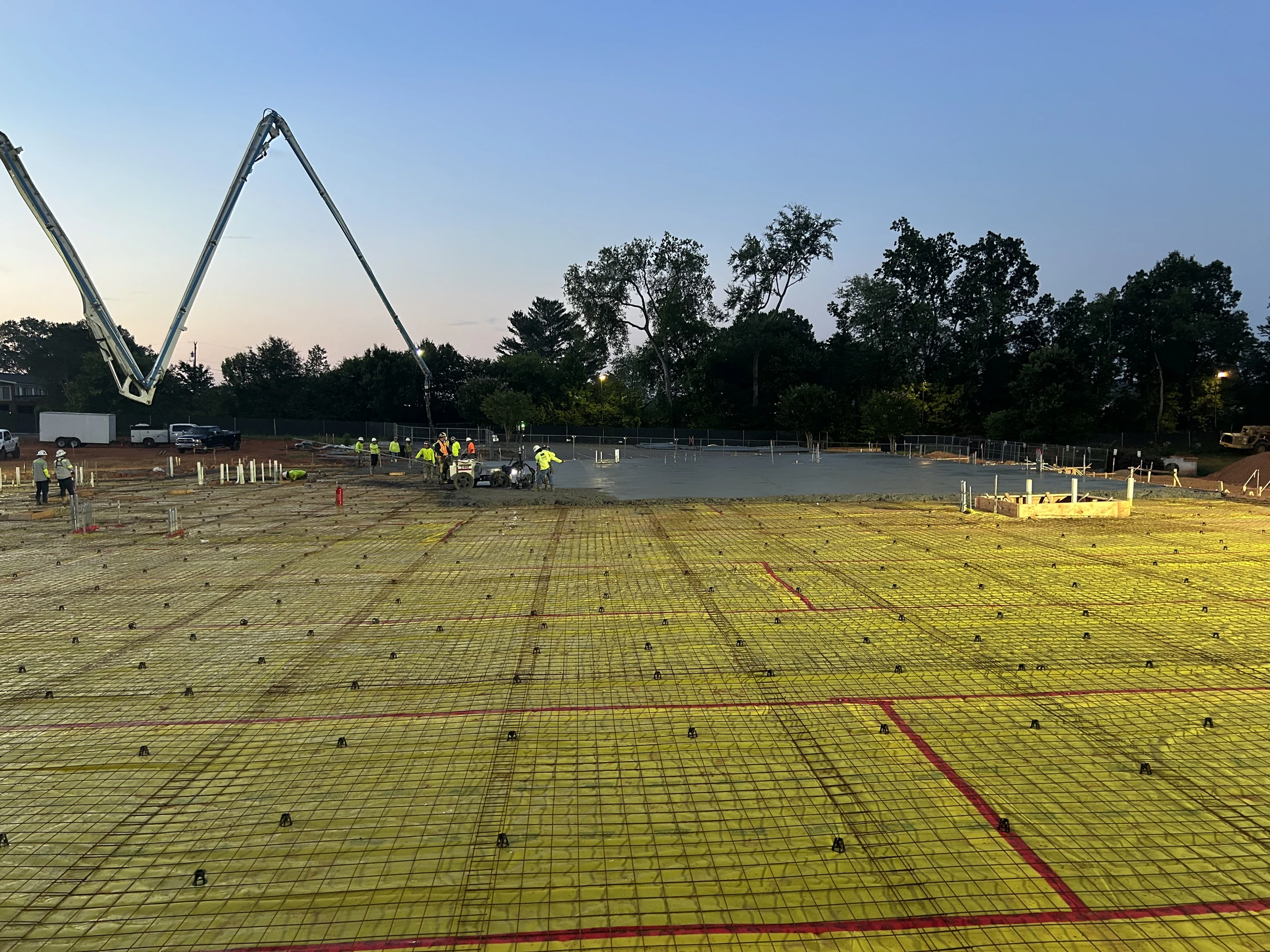 A large construction site at dusk where workers are pouring a concrete slab foundation. A concrete pump truck's boom extends over the work area.