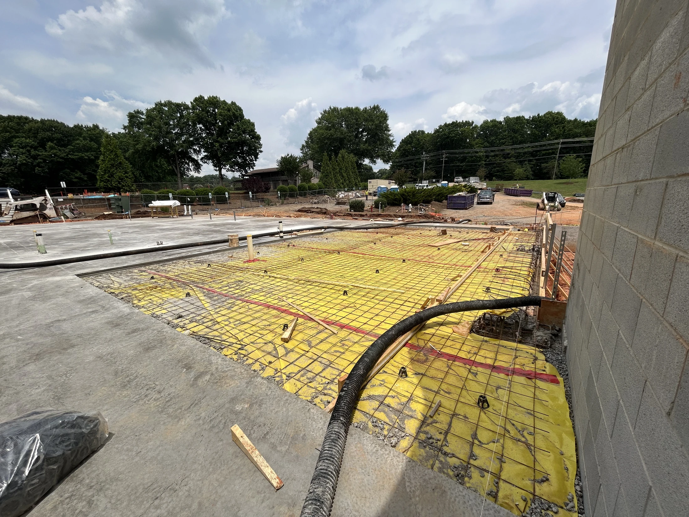 A construction site with a rebar grid set over a yellow vapor barrier, preparing for a concrete pour next to a finished slab and a new block wall.