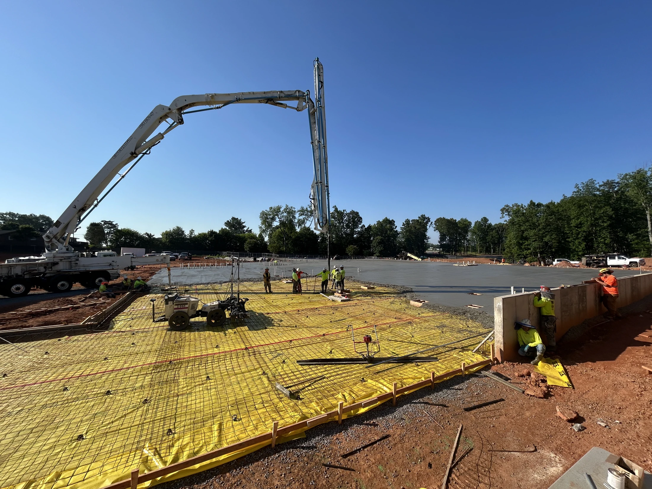 Construction workers using a large pump truck to pour a concrete slab foundation on a sunny day. The area is prepared with a yellow barrier and rebar.