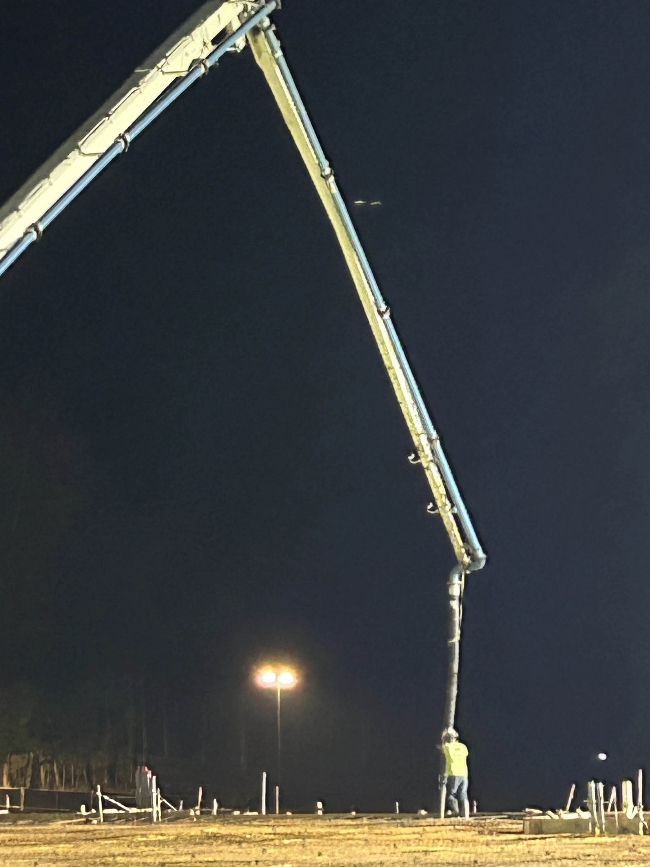 A construction worker in a yellow vest holds a large hose from a concrete pump boom, pouring a foundation at a brightly lit construction site at night.