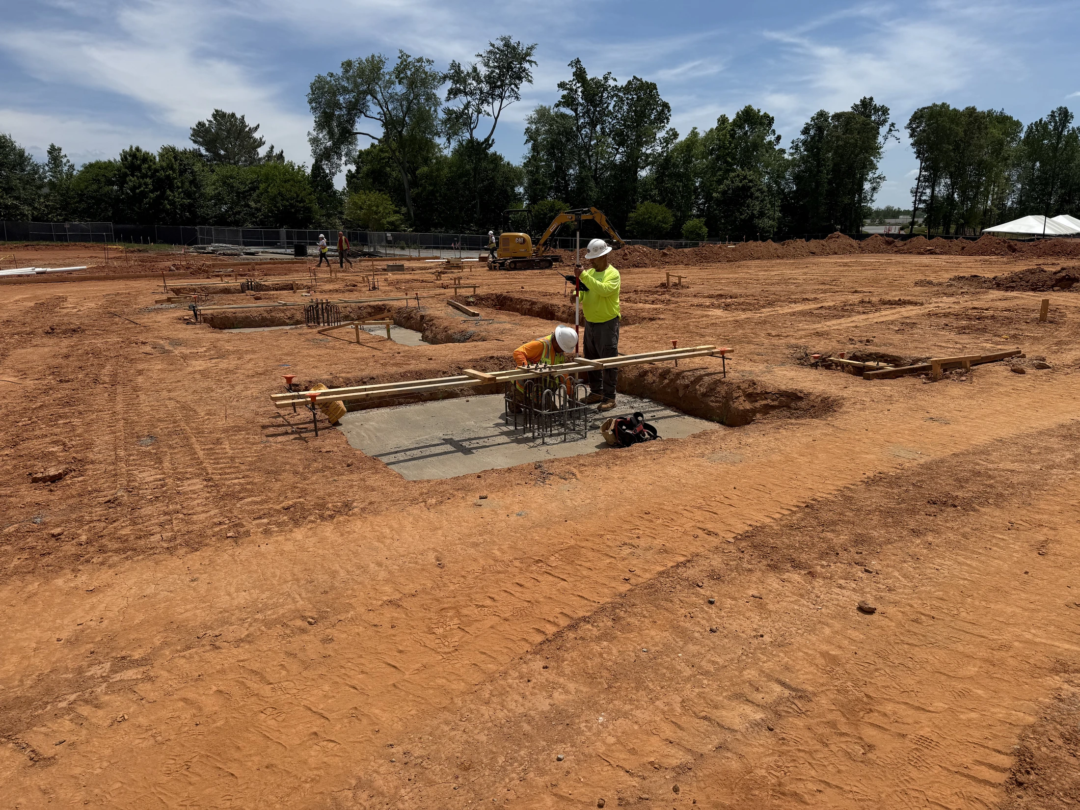 Two workers in hard hats prepare concrete footings with rebar on a large, red dirt construction site, with one worker using surveying equipment.