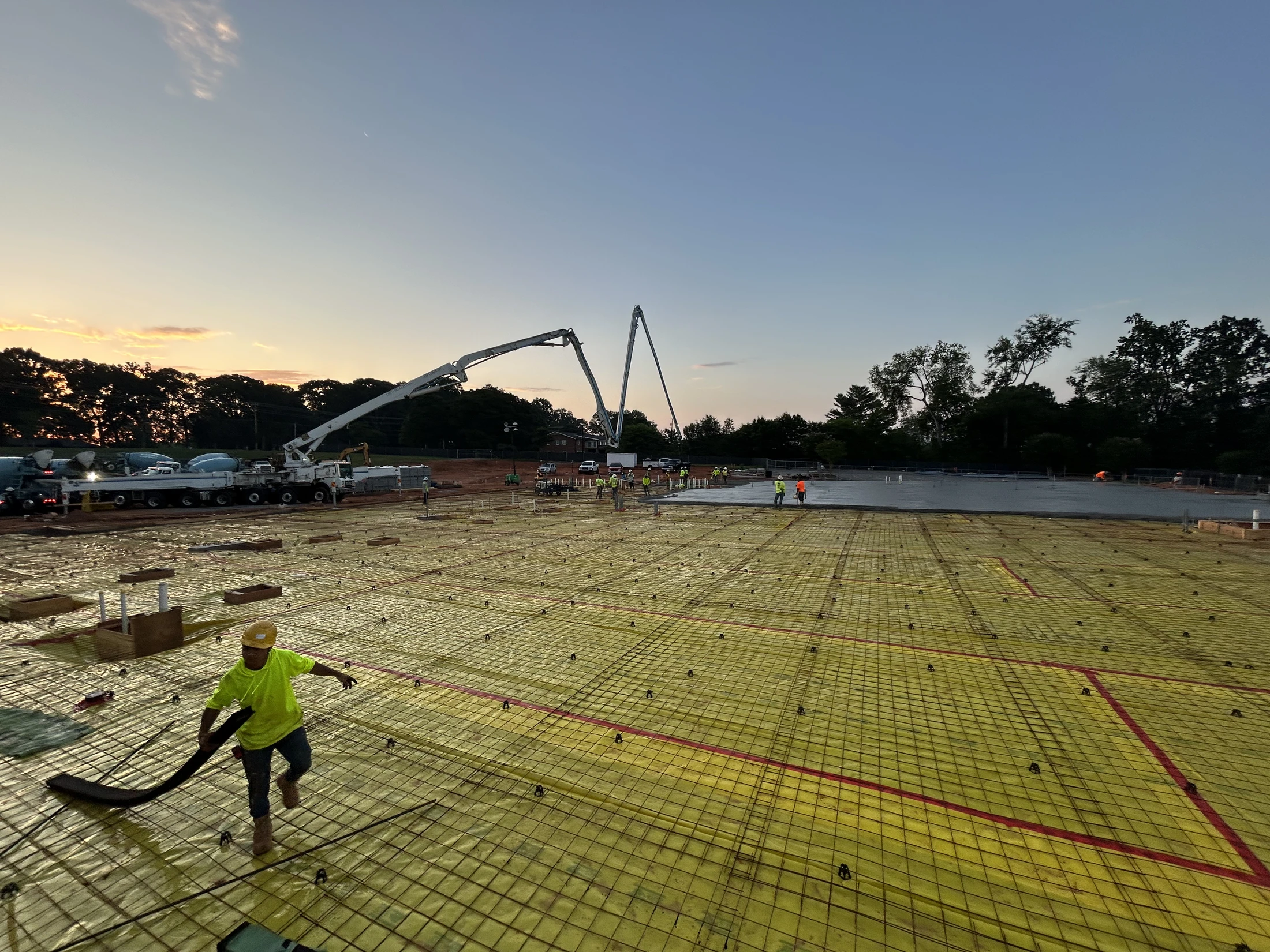 A construction crew works at dawn, pouring a large concrete foundation with a pump truck. A worker walks across a rebar grid in the foreground.