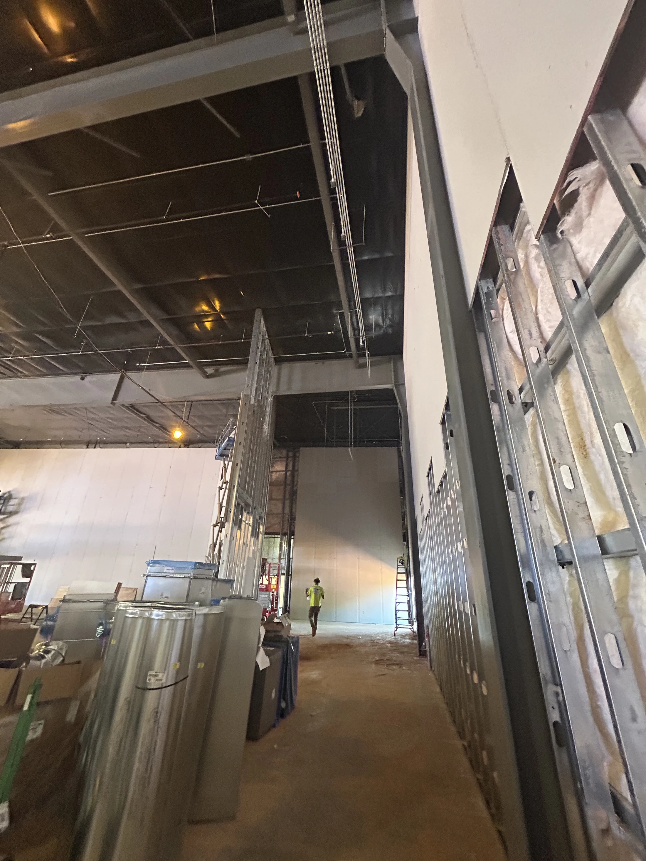 A view down a hallway in a building under construction. Exposed metal wall frames with insulation are on the right, and a worker walks away in the distance.