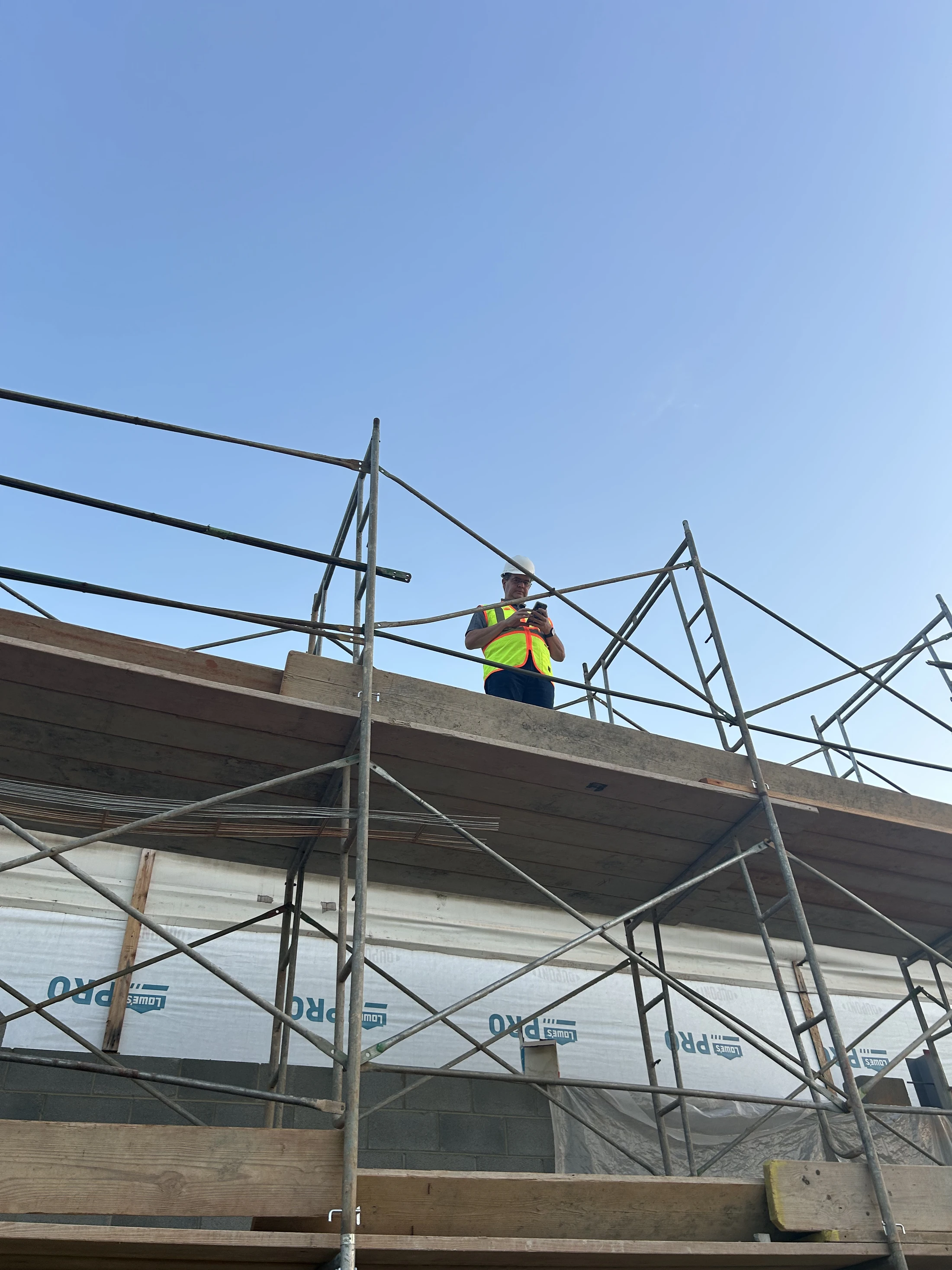 A low-angle view of a construction worker in a hard hat and safety vest, standing on scaffolding and looking at a phone against a clear blue sky.