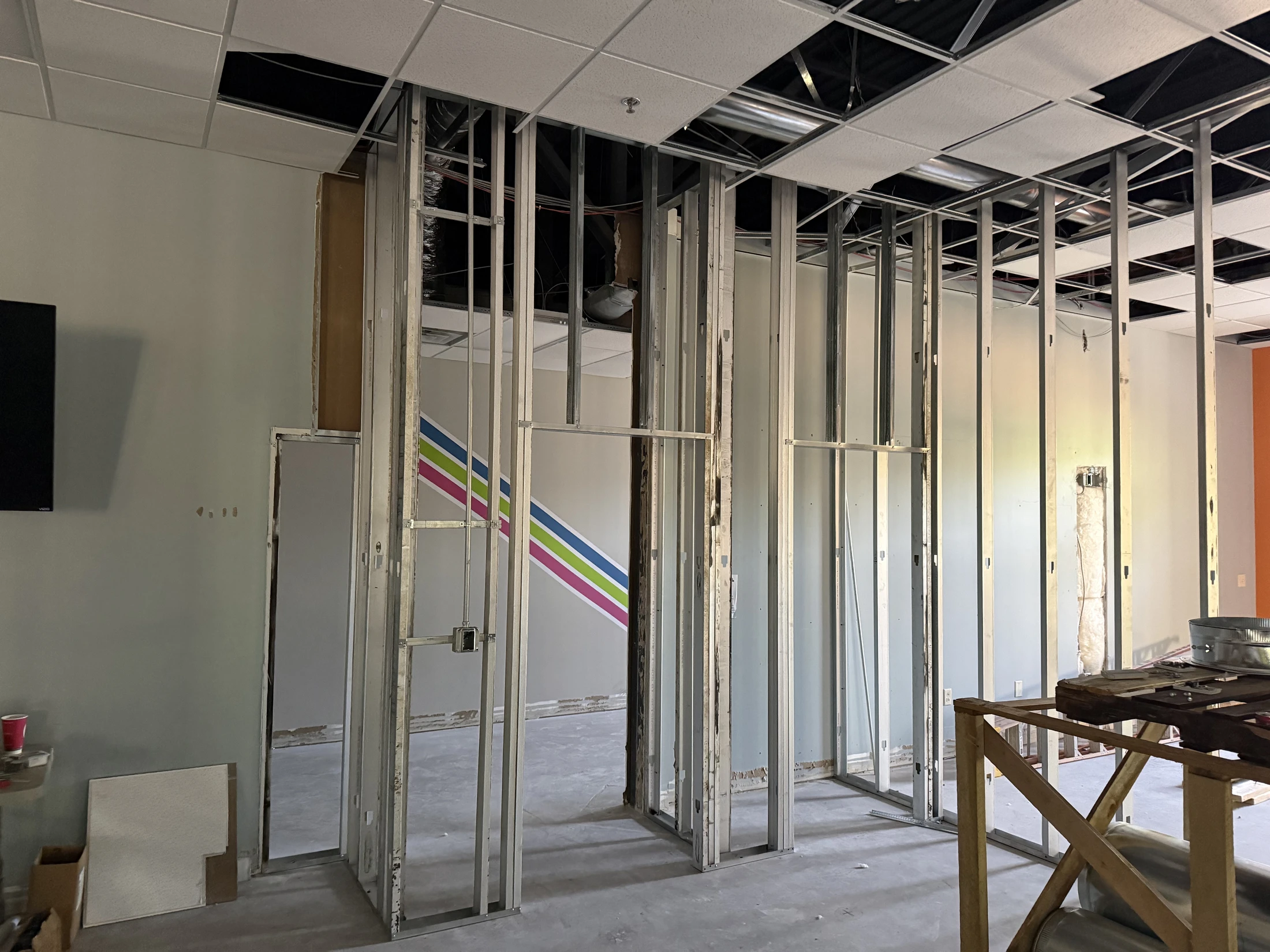 An office interior during renovation, showing exposed metal wall framing, an unfinished drop ceiling, and a concrete floor. A wall has colorful stripes.