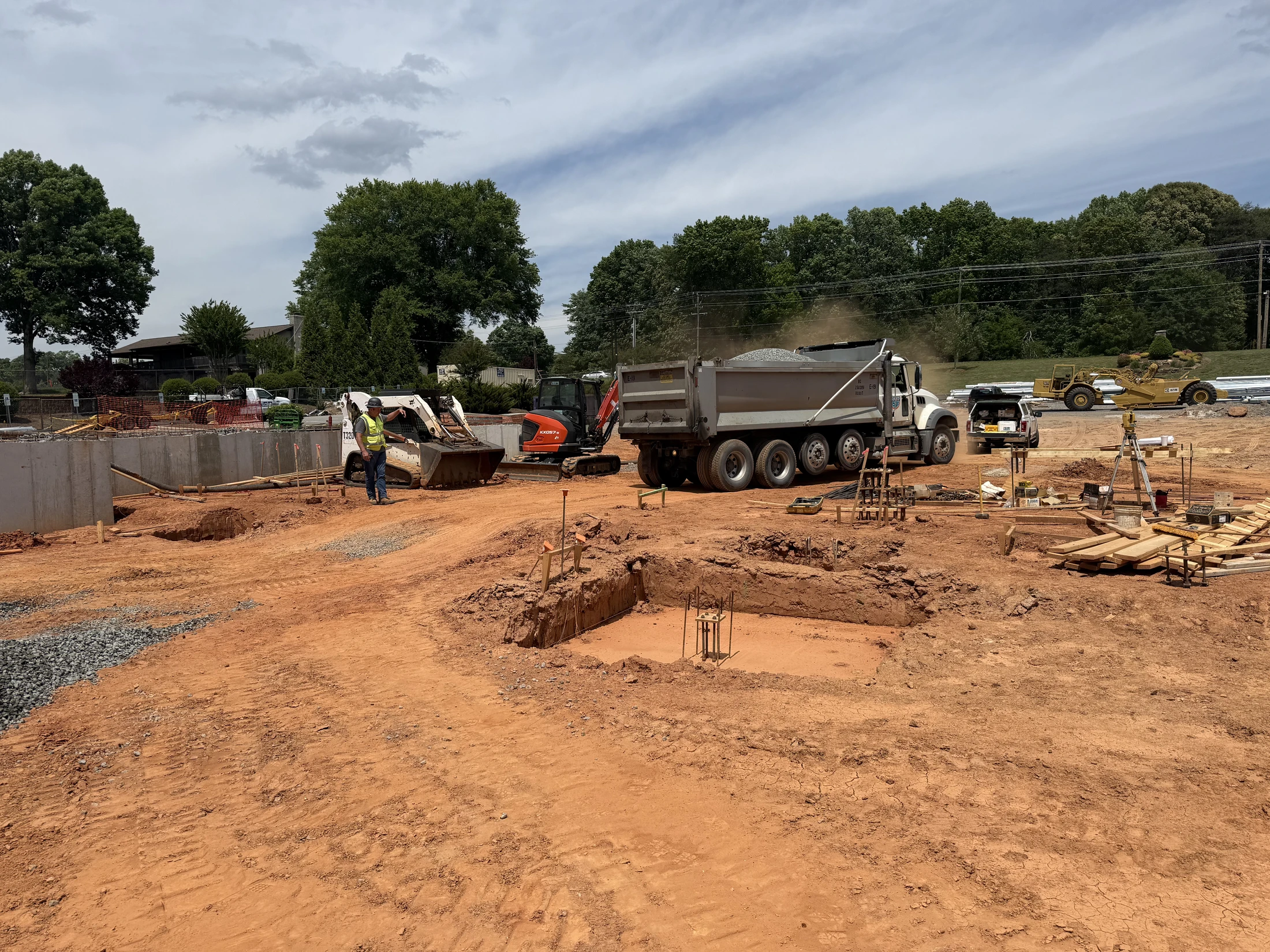 A construction site with red dirt, where a worker stands near a skid steer and mini excavator. A large dump truck is unloading gravel near new foundations.