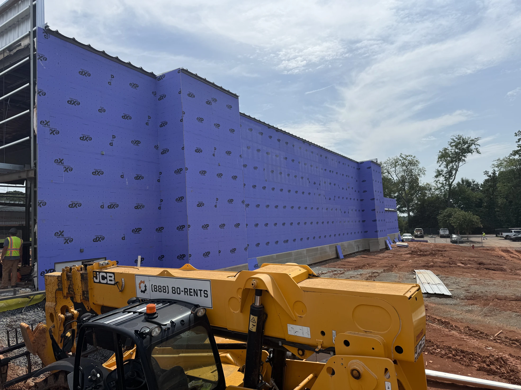A large building under construction with purple exterior sheathing. A yellow JCB telehandler is in the foreground on a dirt lot under a partly cloudy sky.