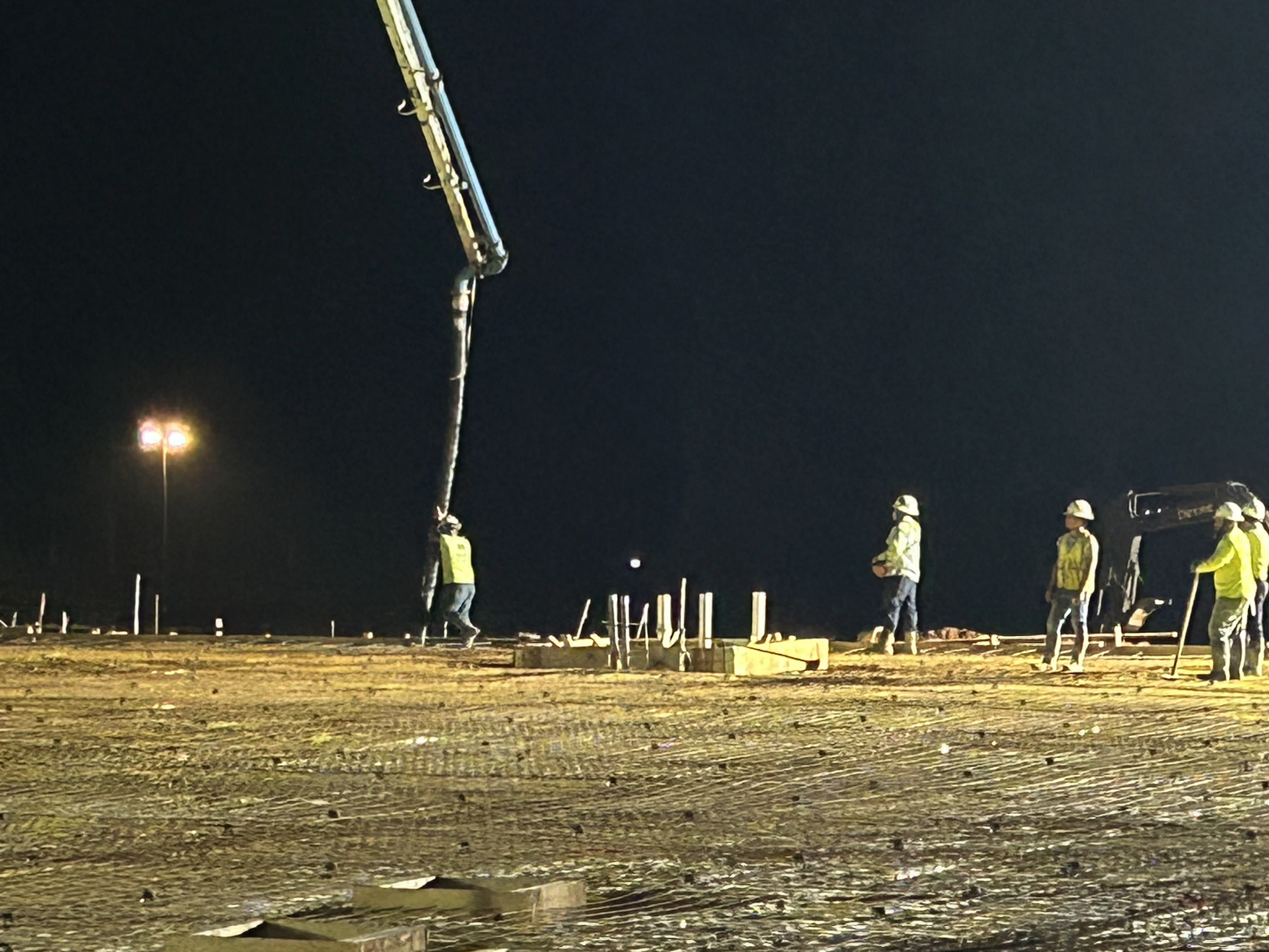 Construction workers pour a concrete slab foundation at night, using a large pump truck to distribute the concrete over a grid of rebar.