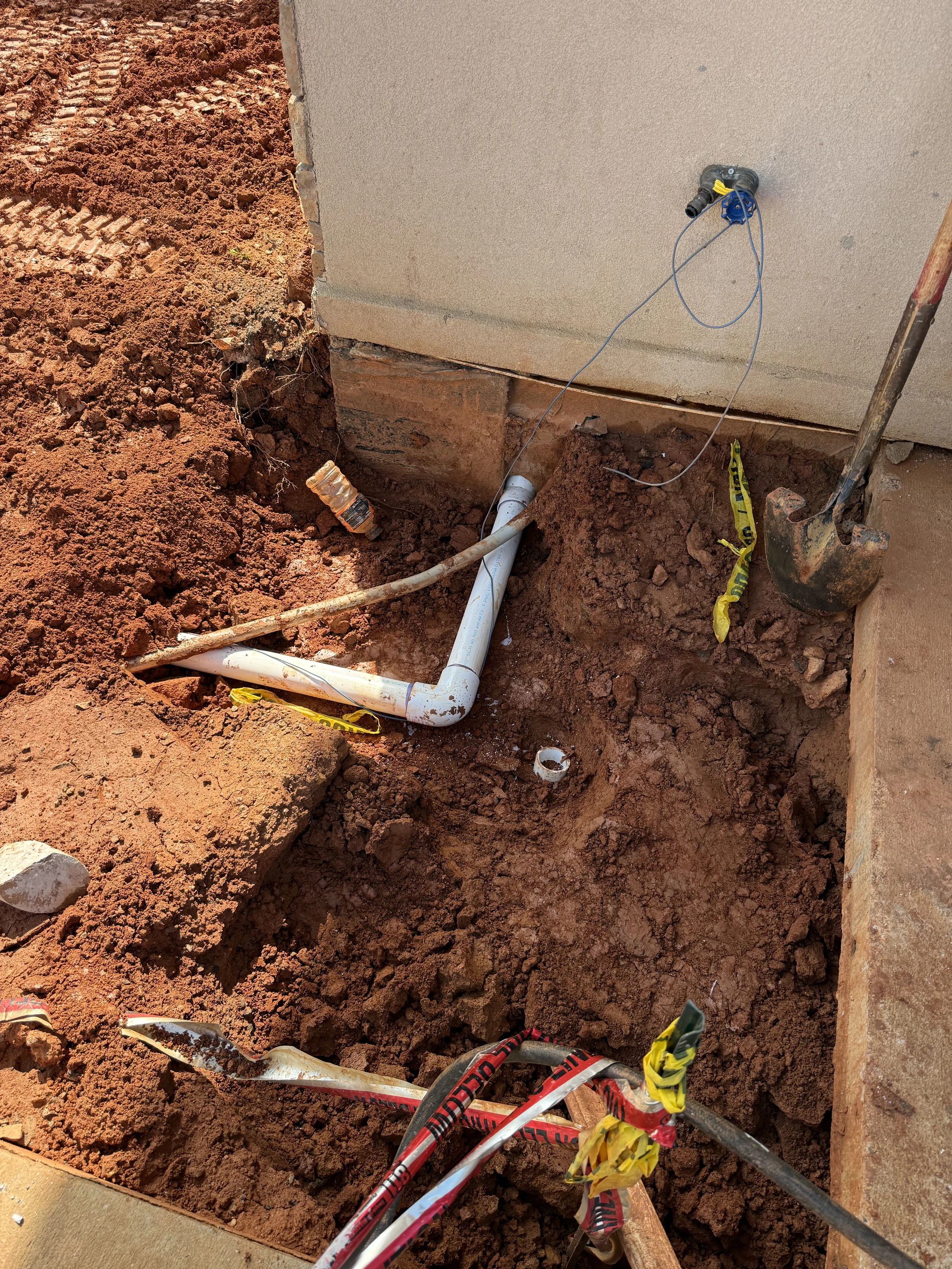 A trench dug in red soil next to a building's foundation exposes white PVC pipes and utility lines. A shovel leans against the concrete nearby.