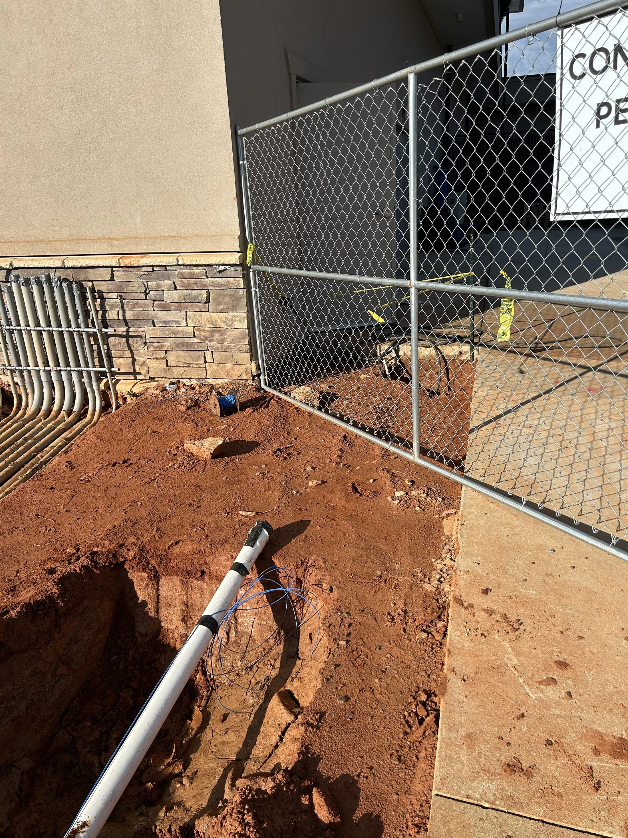 A construction site with a trench dug in red clay next to a building. A white pipe and wires are exposed, with a chain-link fence in the background.