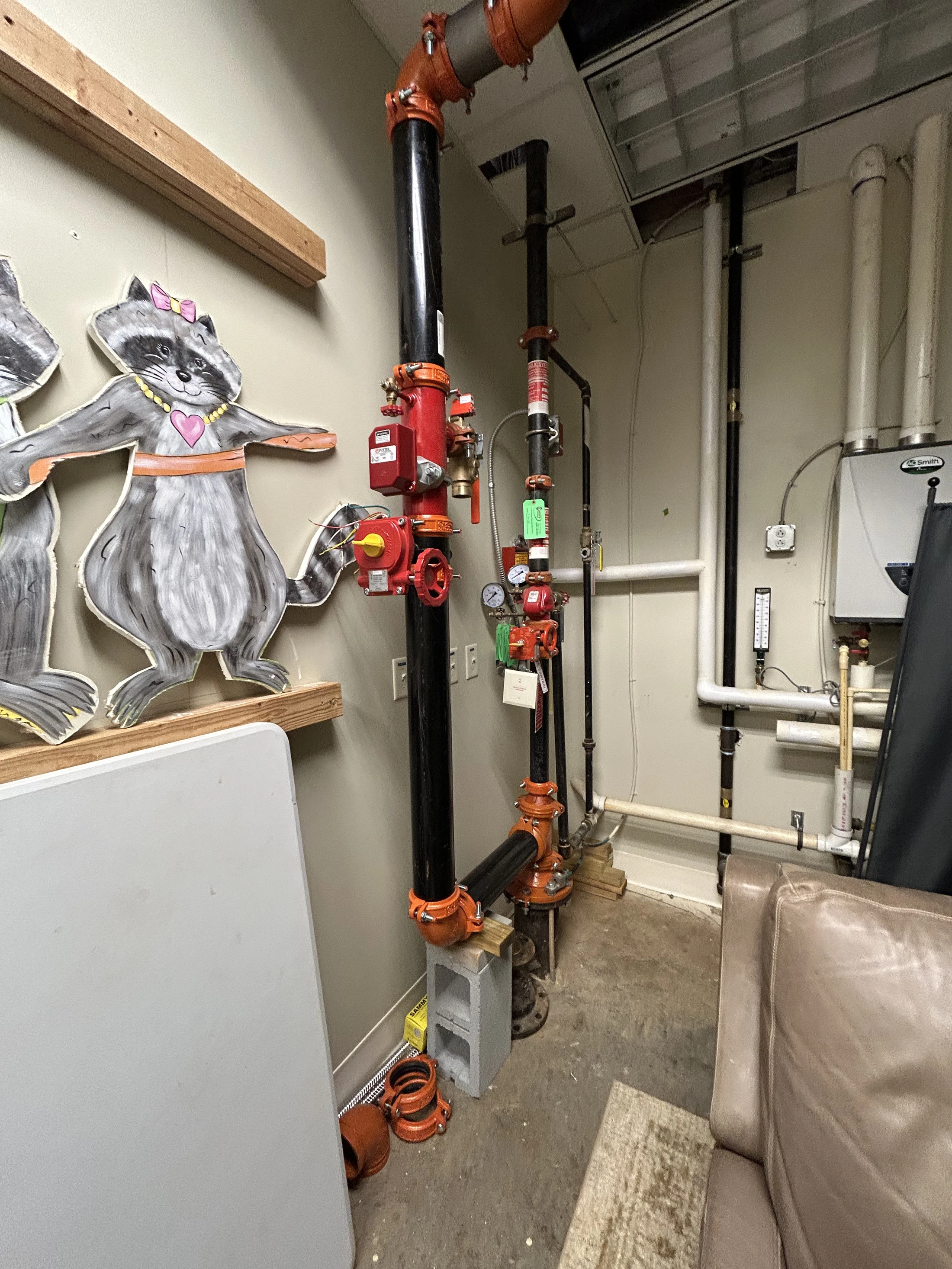 A utility room with large black and red fire sprinkler pipes next to painted cardboard cutouts of cartoon raccoons resting on wooden shelves.