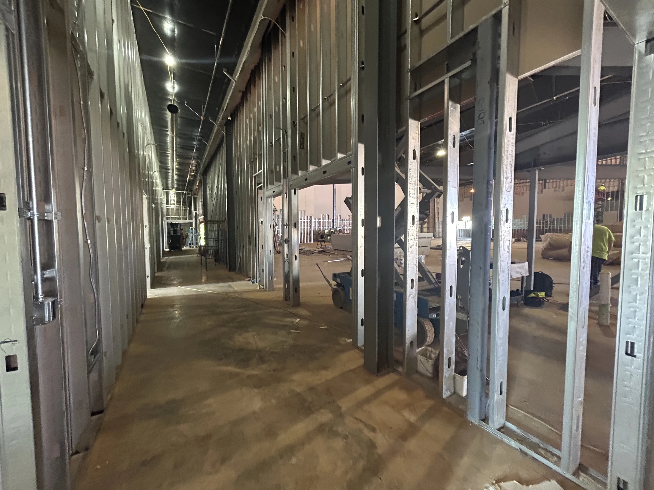 A long hallway in a building under construction. The walls are exposed metal studs and the floor is concrete. Temporary lights illuminate the space.