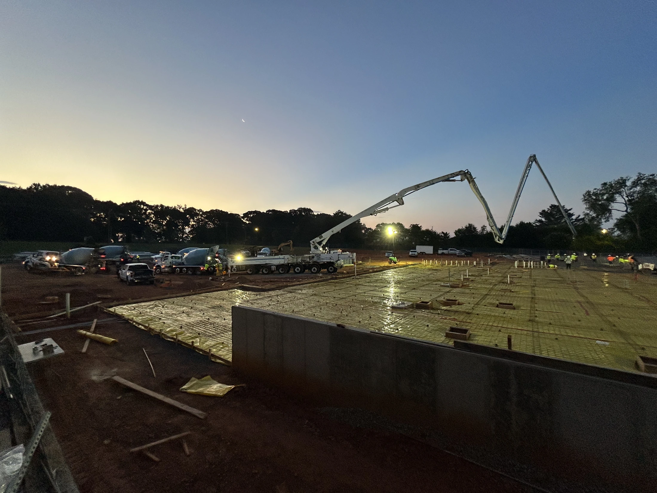 A large construction site at dusk or dawn, where a concrete pump truck is pouring a foundation slab over a grid of rebar, with mixer trucks lined up.
