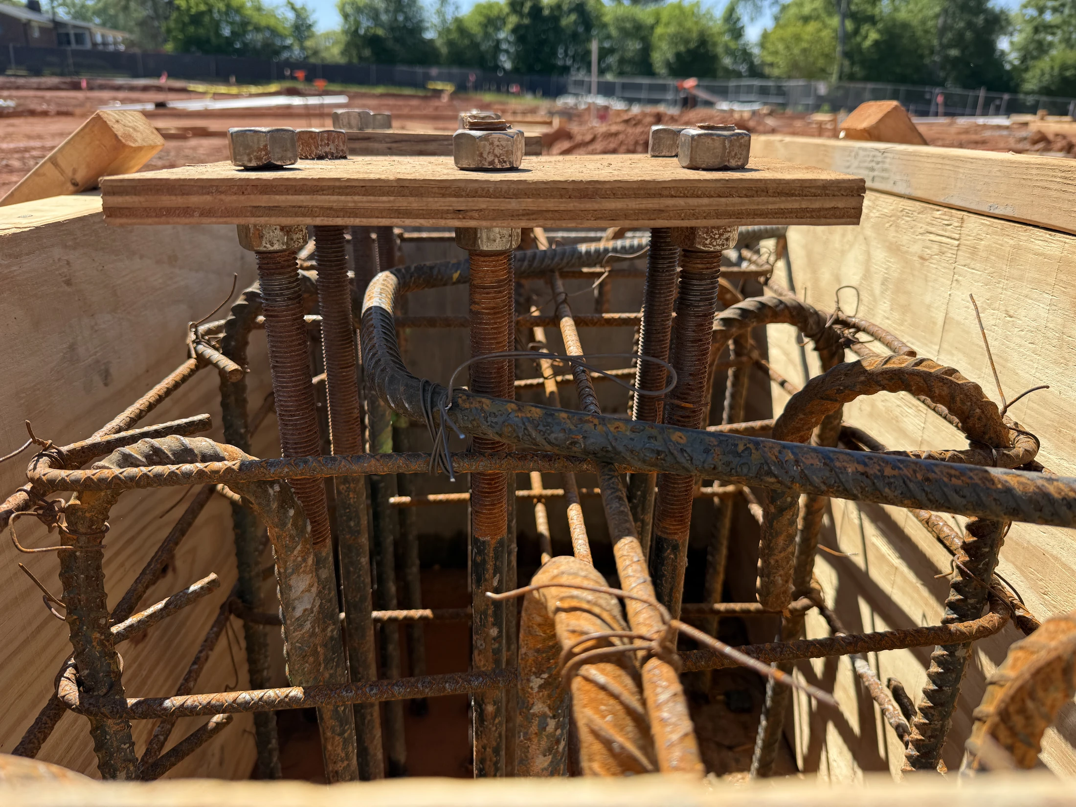A close-up shot inside a wooden concrete form, showing a cage of rusty rebar and large anchor bolts held in place by a wood template and nuts.
