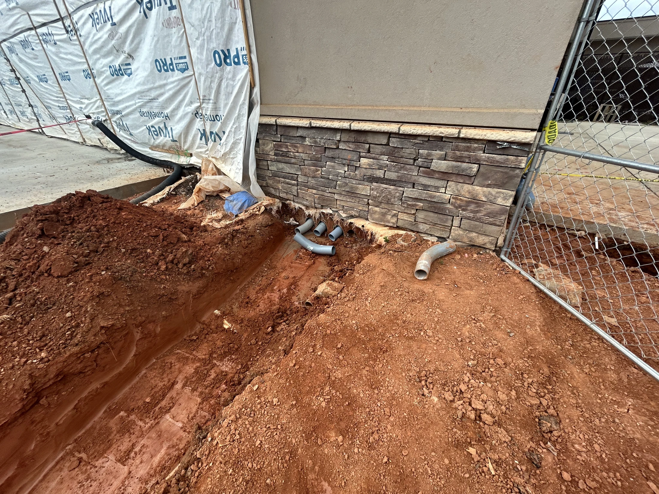 A construction site with a trench dug in red soil next to a building's stone foundation, revealing several sections of grey PVC piping.