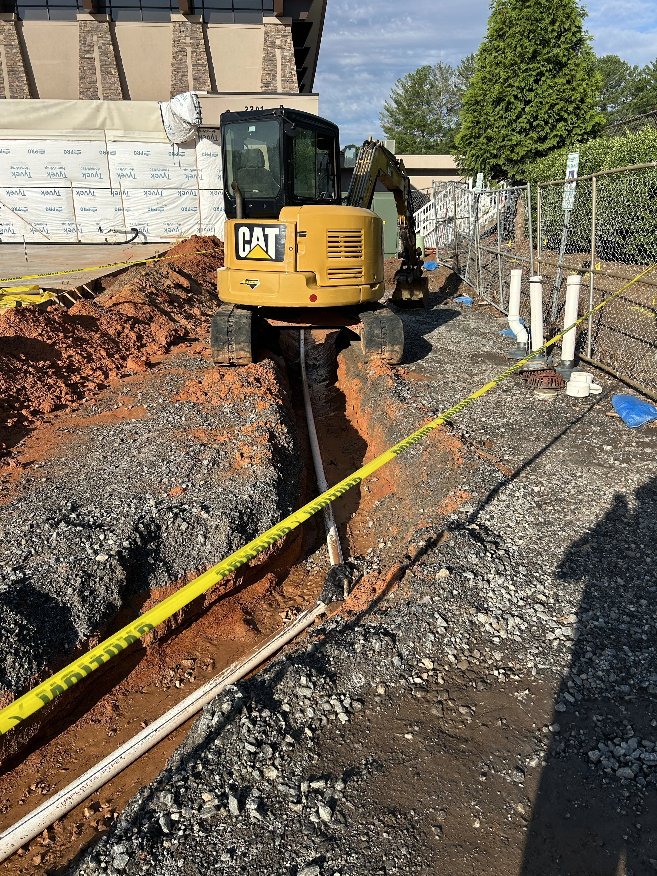 A yellow CAT mini excavator straddles a narrow trench dug in red clay and gravel. White PVC pipes are visible inside the trench at a construction site.