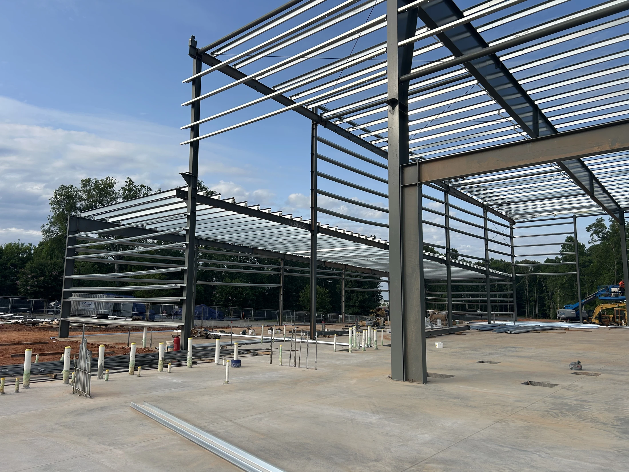 The steel framework of a large new building rises from a concrete foundation on a construction site. White pipes for plumbing emerge from the concrete floor.