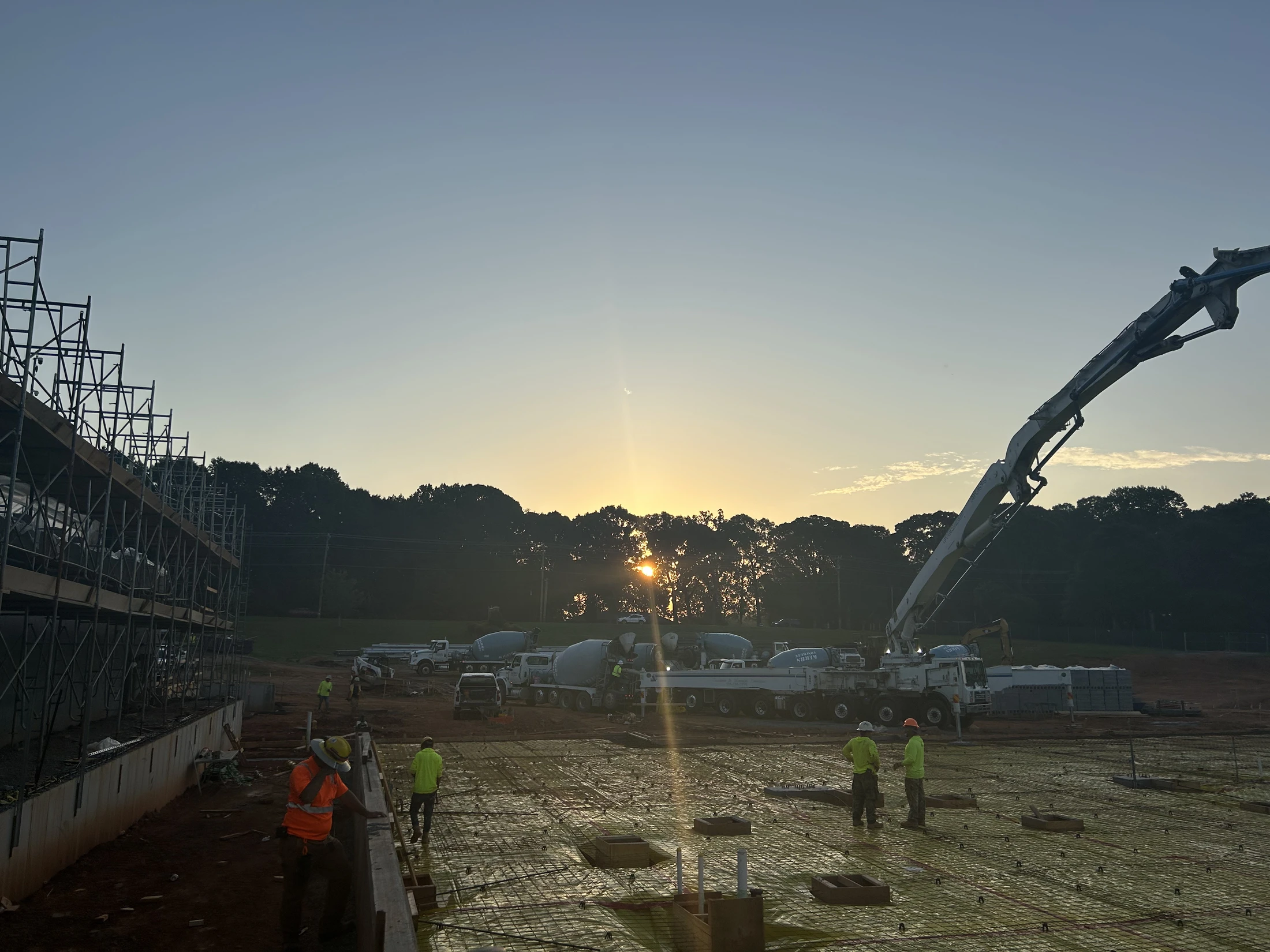 A construction site at sunrise where workers are preparing to pour a concrete foundation. A concrete pump truck and mixer trucks are ready for the pour.