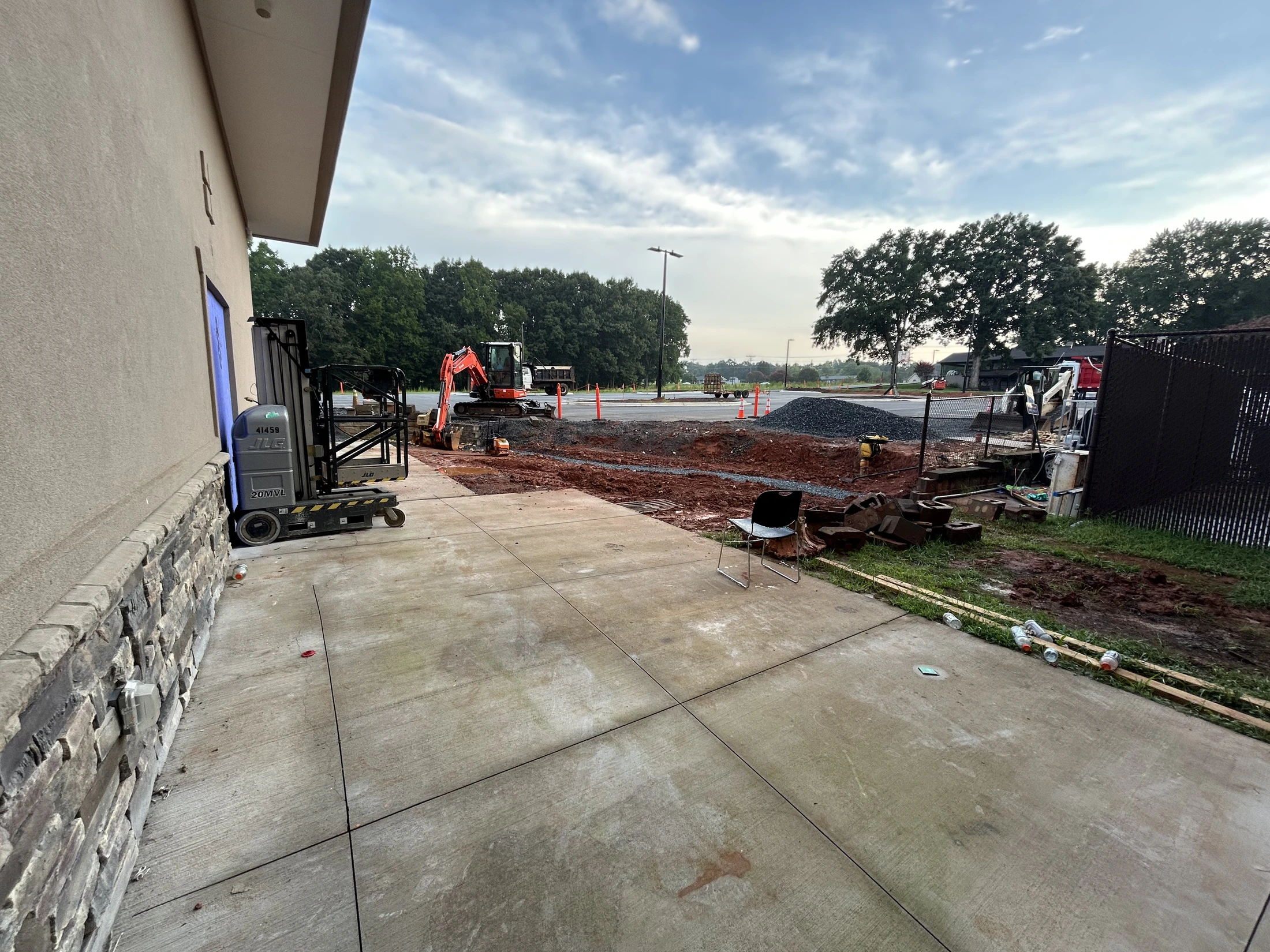 A construction site next to a building. The foreground shows a concrete patio, while the background has an excavator, dirt, and a pile of gravel.