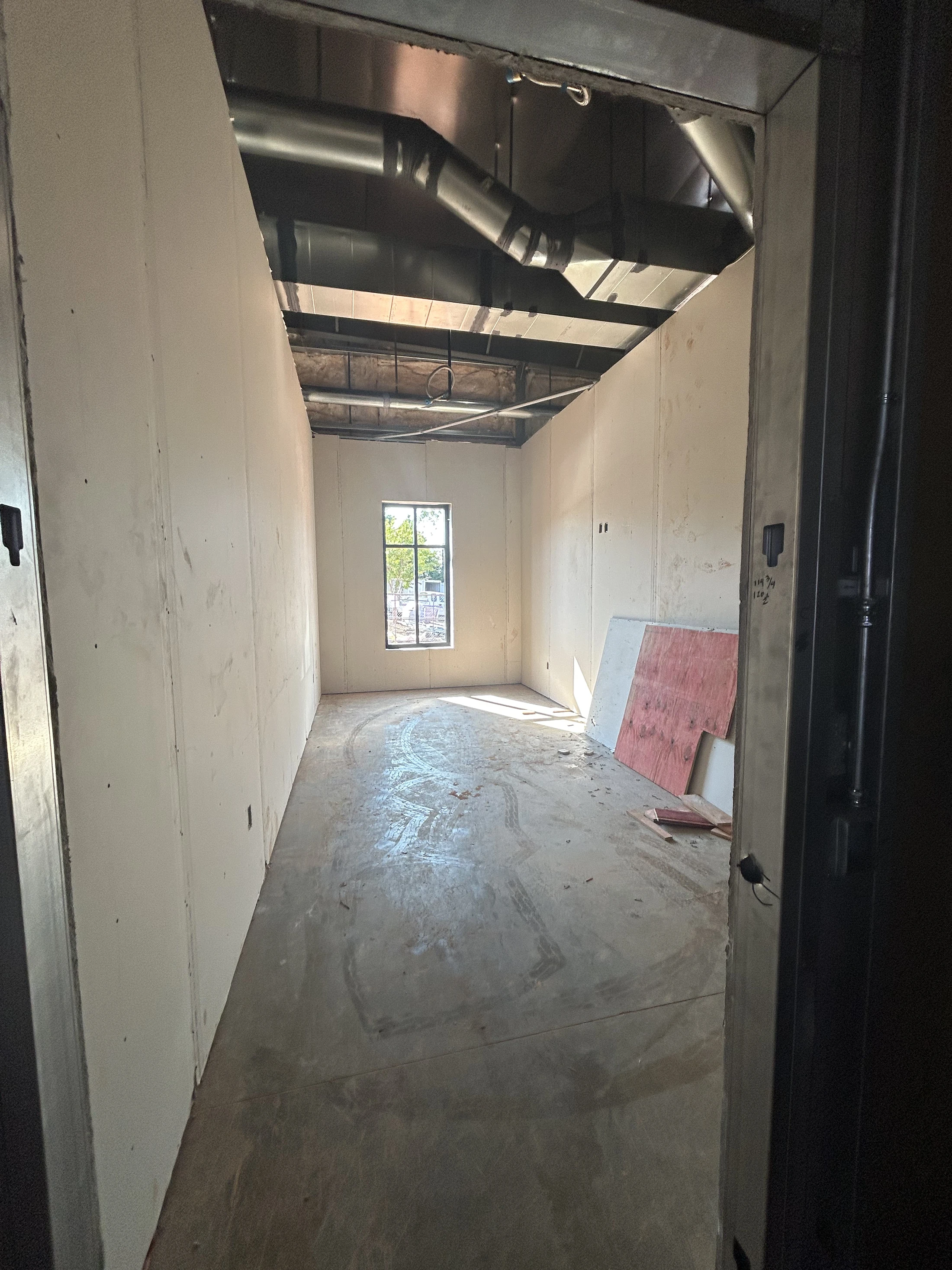 A view from a doorway into an empty, unfinished room with drywall, a concrete floor with tire tracks, and exposed ductwork on the ceiling.