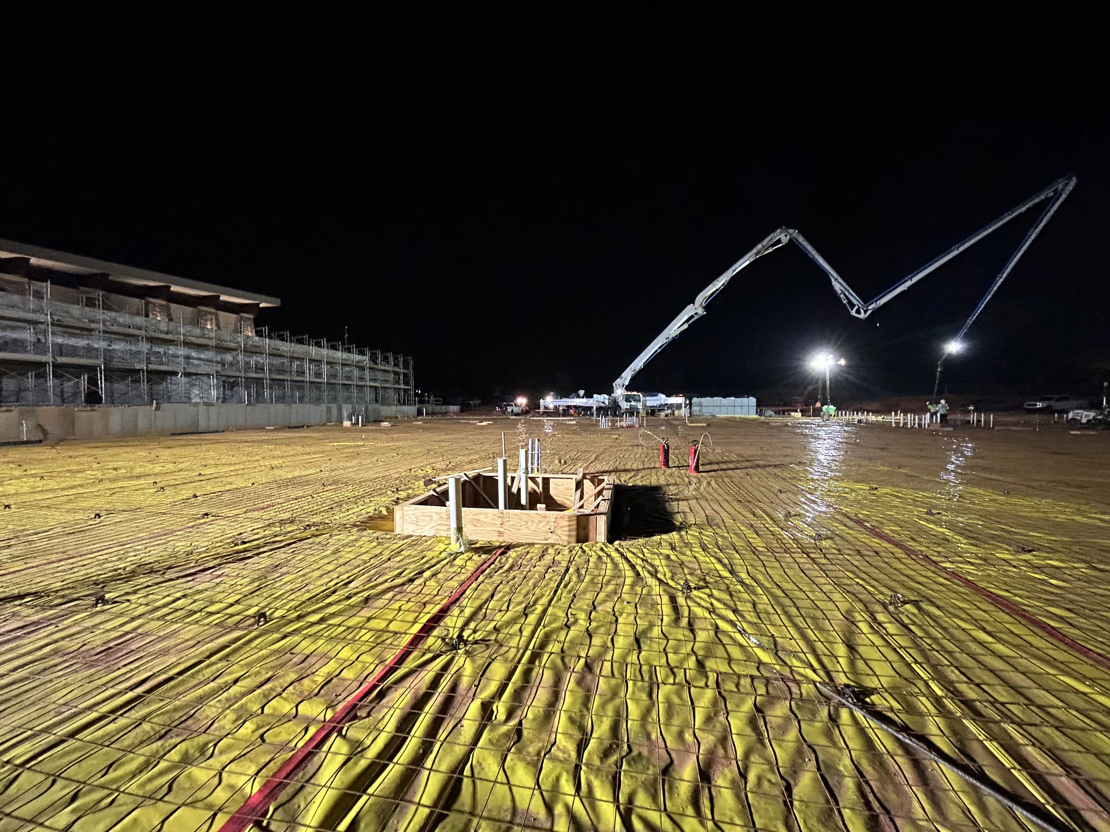 A construction site at night, prepped for a large concrete slab pour. A concrete pump truck's boom extends over a rebar grid on a yellow vapor barrier.