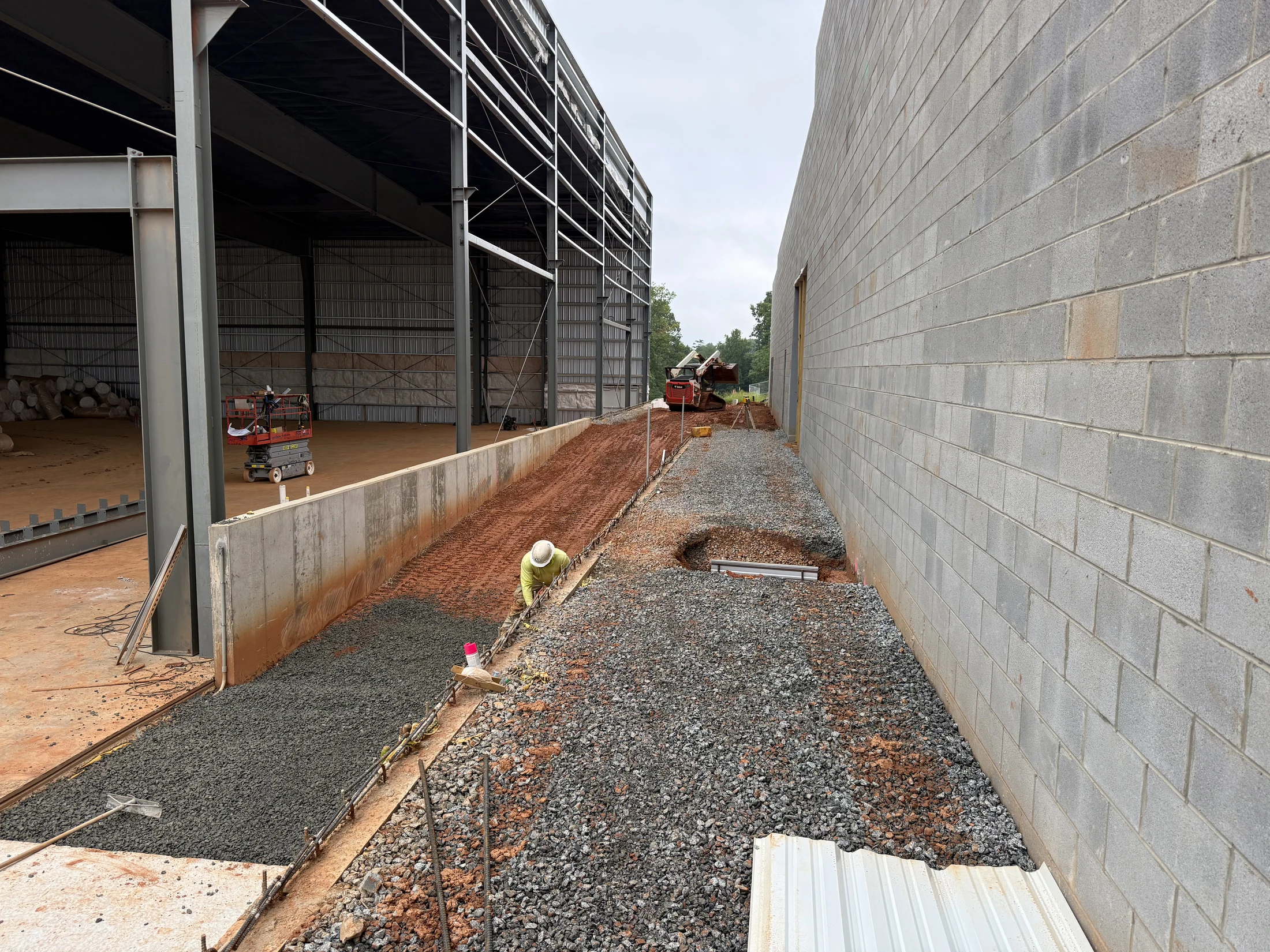 A worker in a hard hat prepares rebar for a concrete pour between a steel-framed building and a block wall at a construction site.