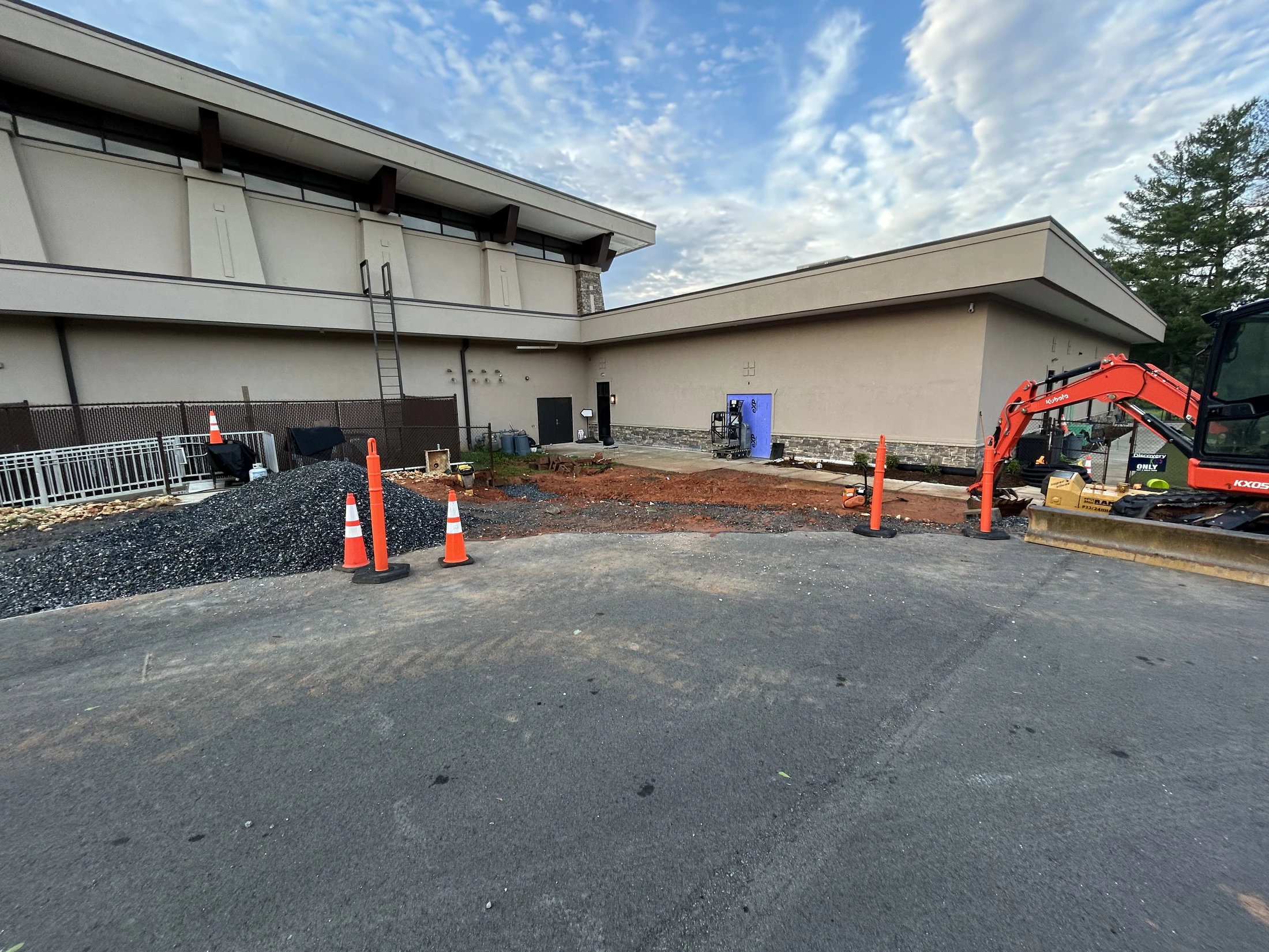 The exterior of a building undergoing renovation, showing an orange excavator, dirt, gravel, and traffic cones on a paved surface.