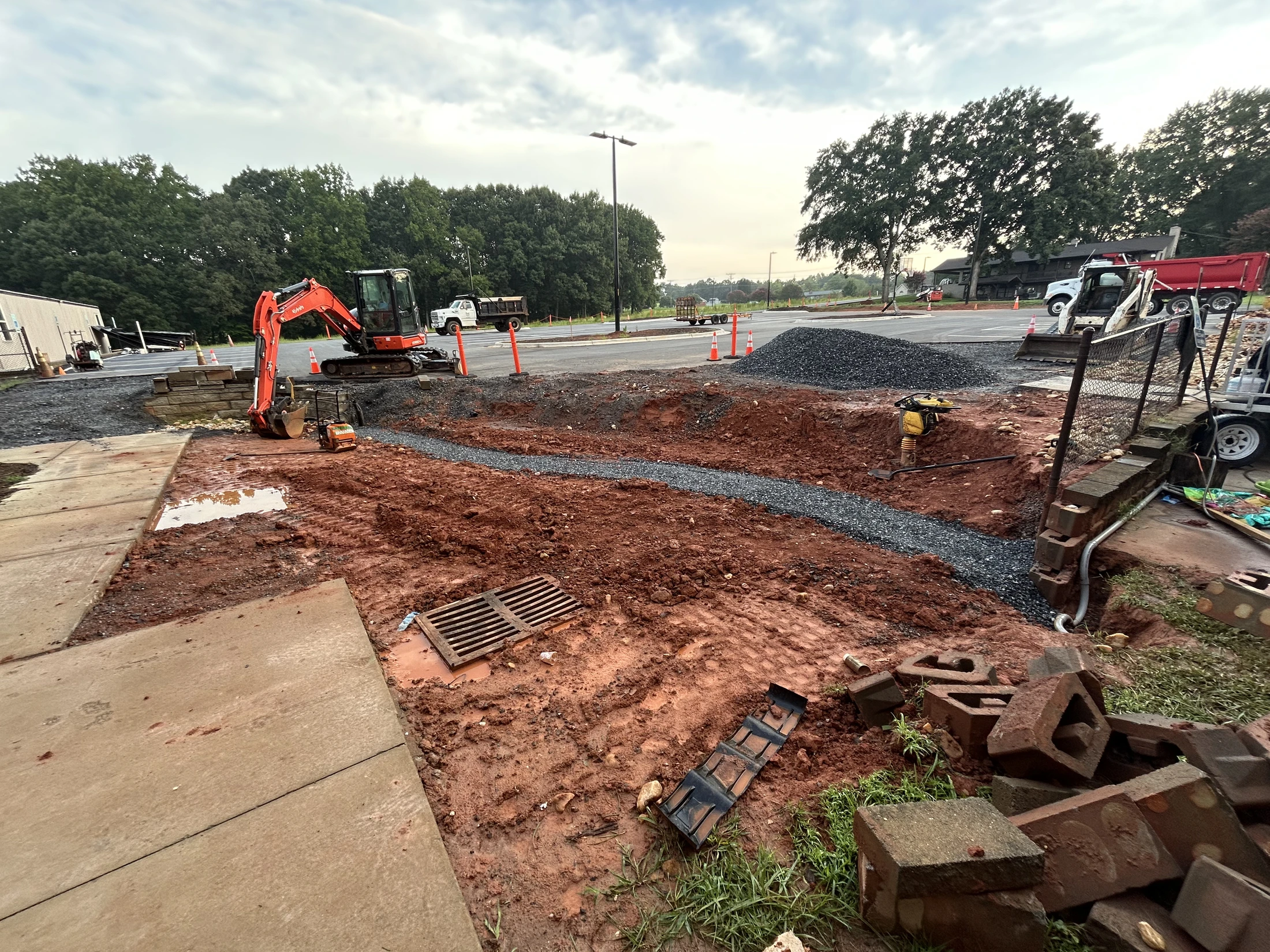 A construction site with an orange excavator on red clay soil. A newly dug trench filled with gray gravel snakes through the muddy ground.