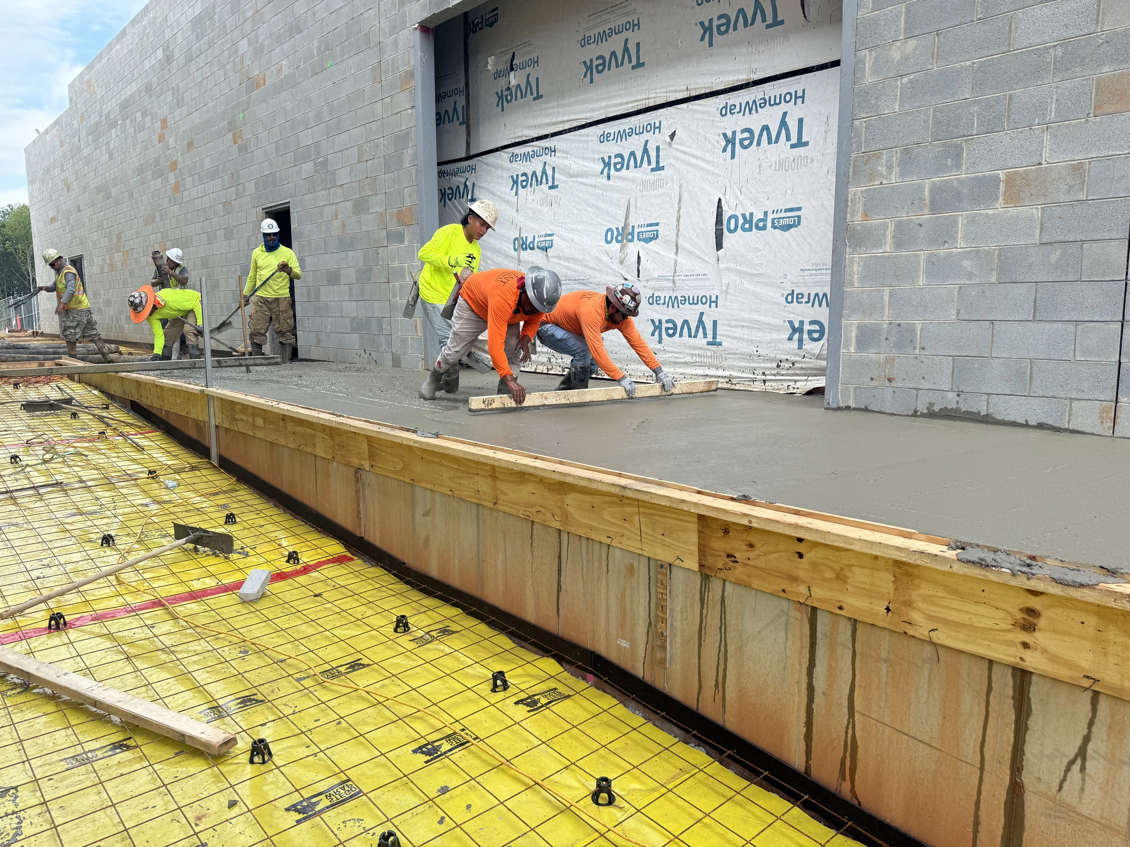 A team of construction workers in hard hats and safety gear smooths freshly poured wet concrete next to a cinder block building under construction.