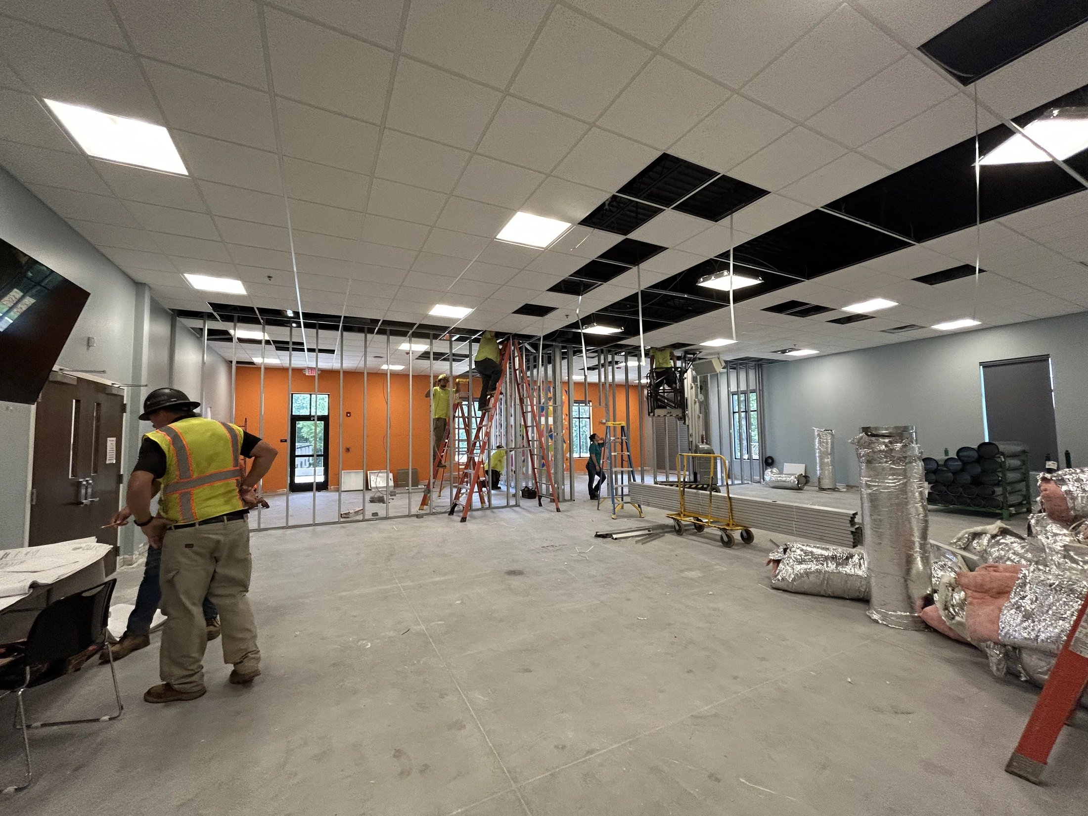 A wide view of a commercial interior under construction, with several workers on ladders installing ceiling tiles and building a metal stud wall.