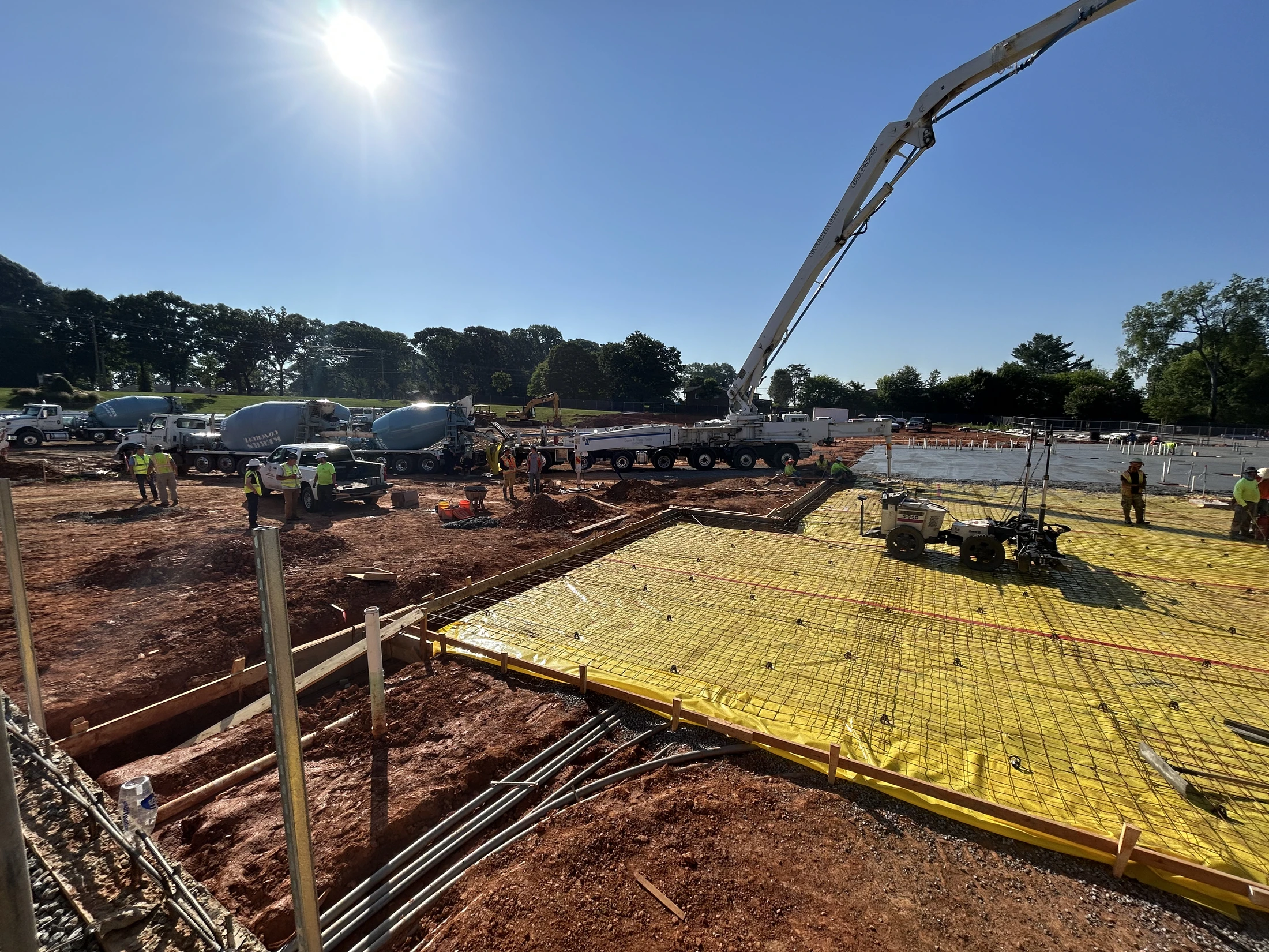 A large construction site on a sunny day, with cement trucks and a pump truck pouring a concrete foundation over a yellow vapor barrier and rebar grid.
