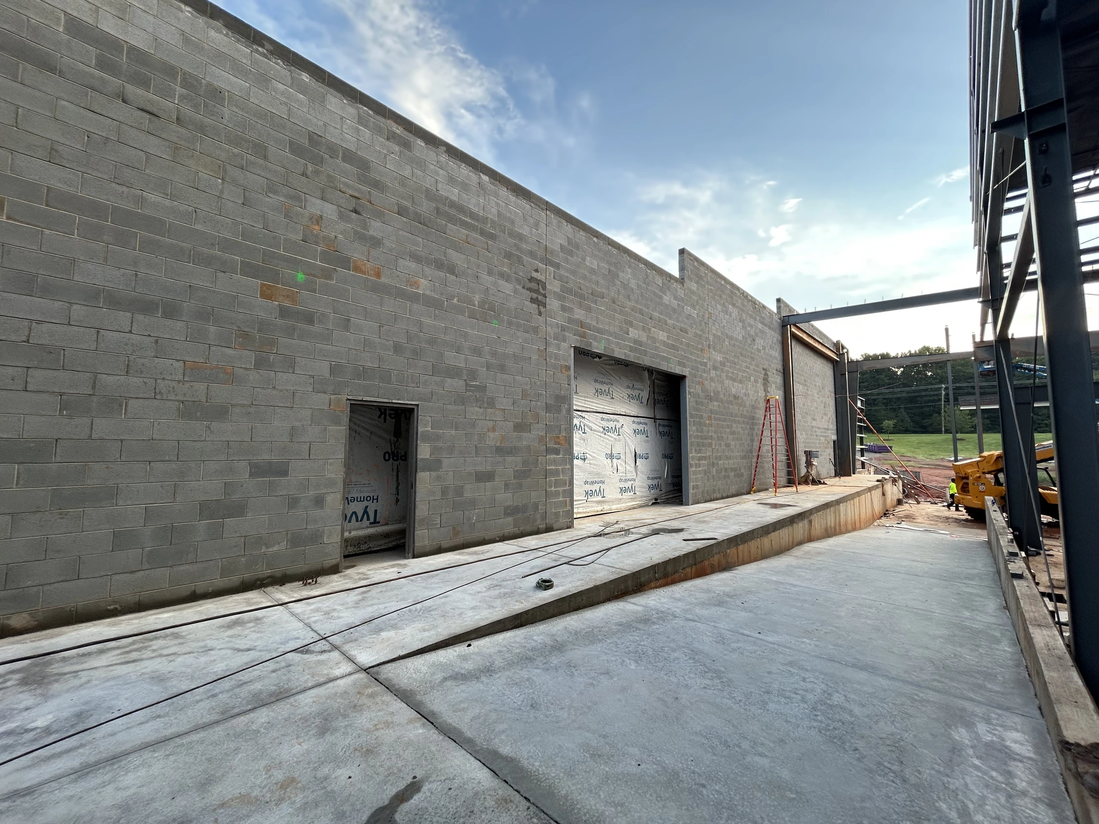 A long grey concrete block wall of a building under construction, with a concrete loading dock and ramp in front and steel framing visible on the right.