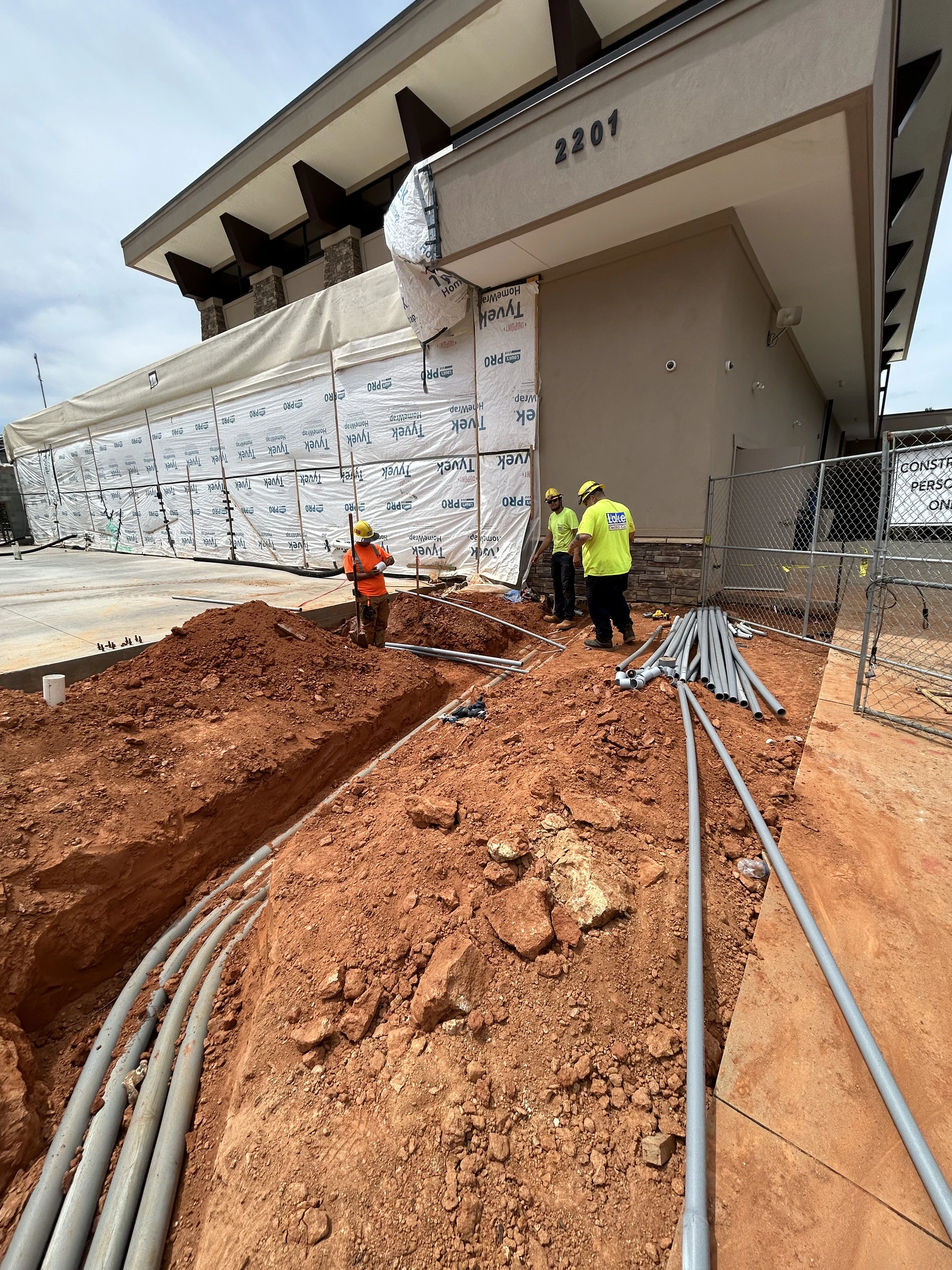 Three construction workers install grey electrical conduits in a trench dug in red dirt next to a commercial building that is under construction.