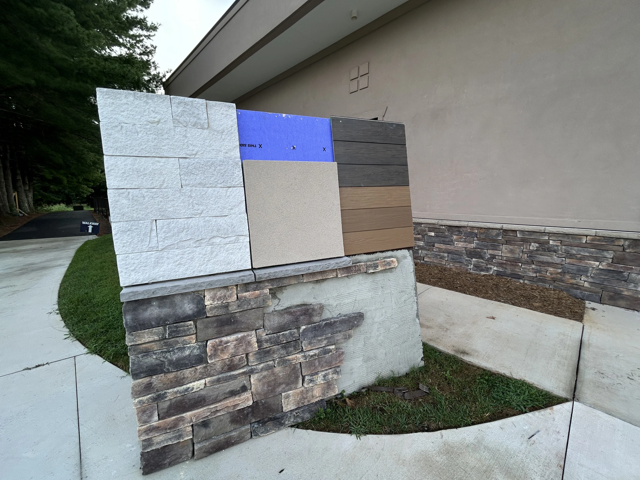 An outdoor sample wall showing various construction materials, including a stacked stone base, white stone panels, stucco, and two colors of wood-look siding.