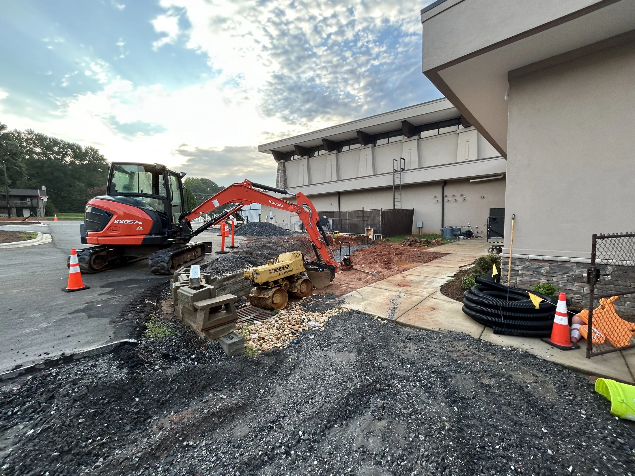 An orange excavator and a small yellow trench roller parked at a construction site next to a building, with piles of gravel and dirt on the ground.