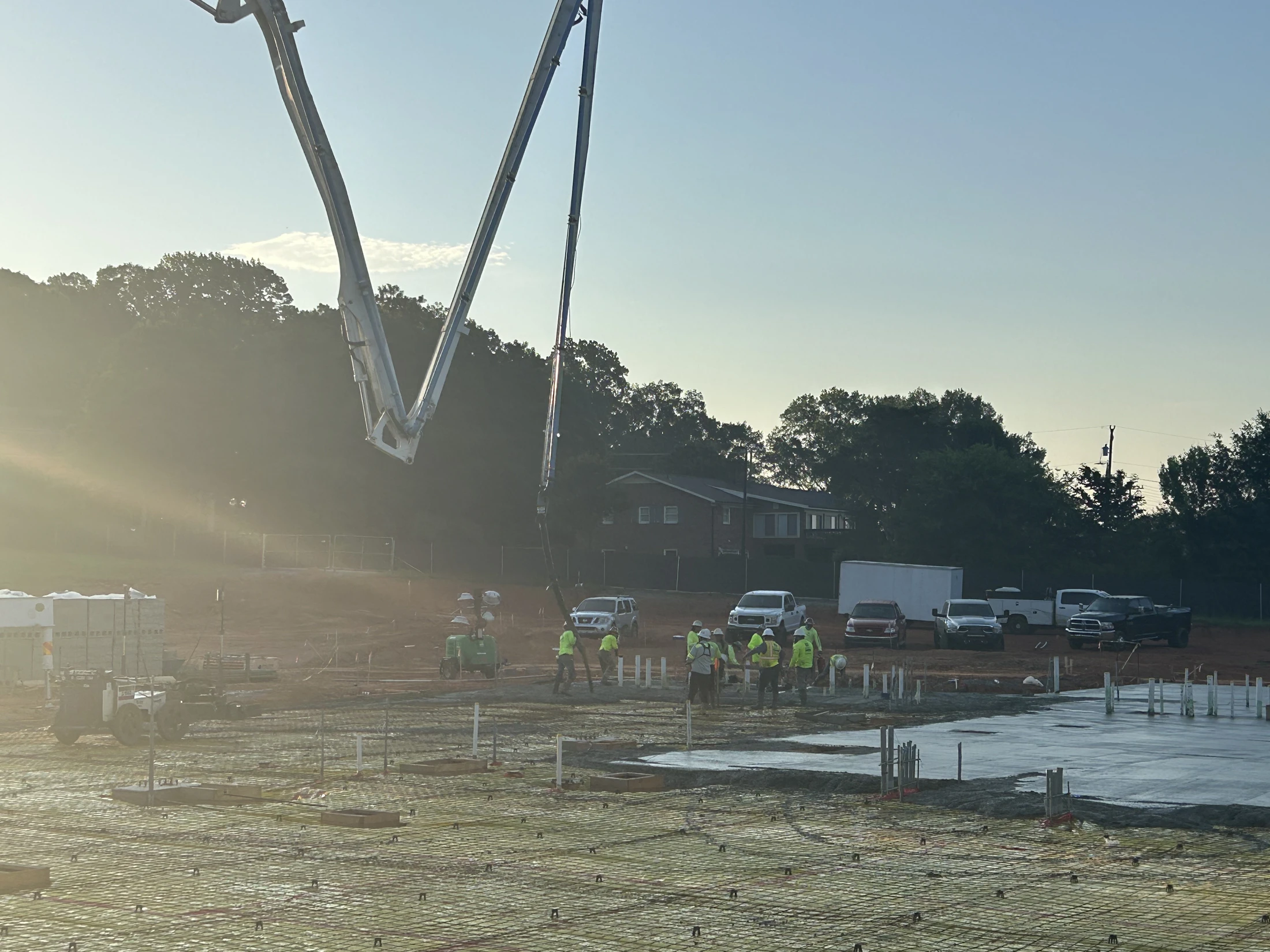 A construction crew uses a large concrete pump truck to pour a foundation slab on a sunny morning. Workers in hard hats guide the concrete into place.