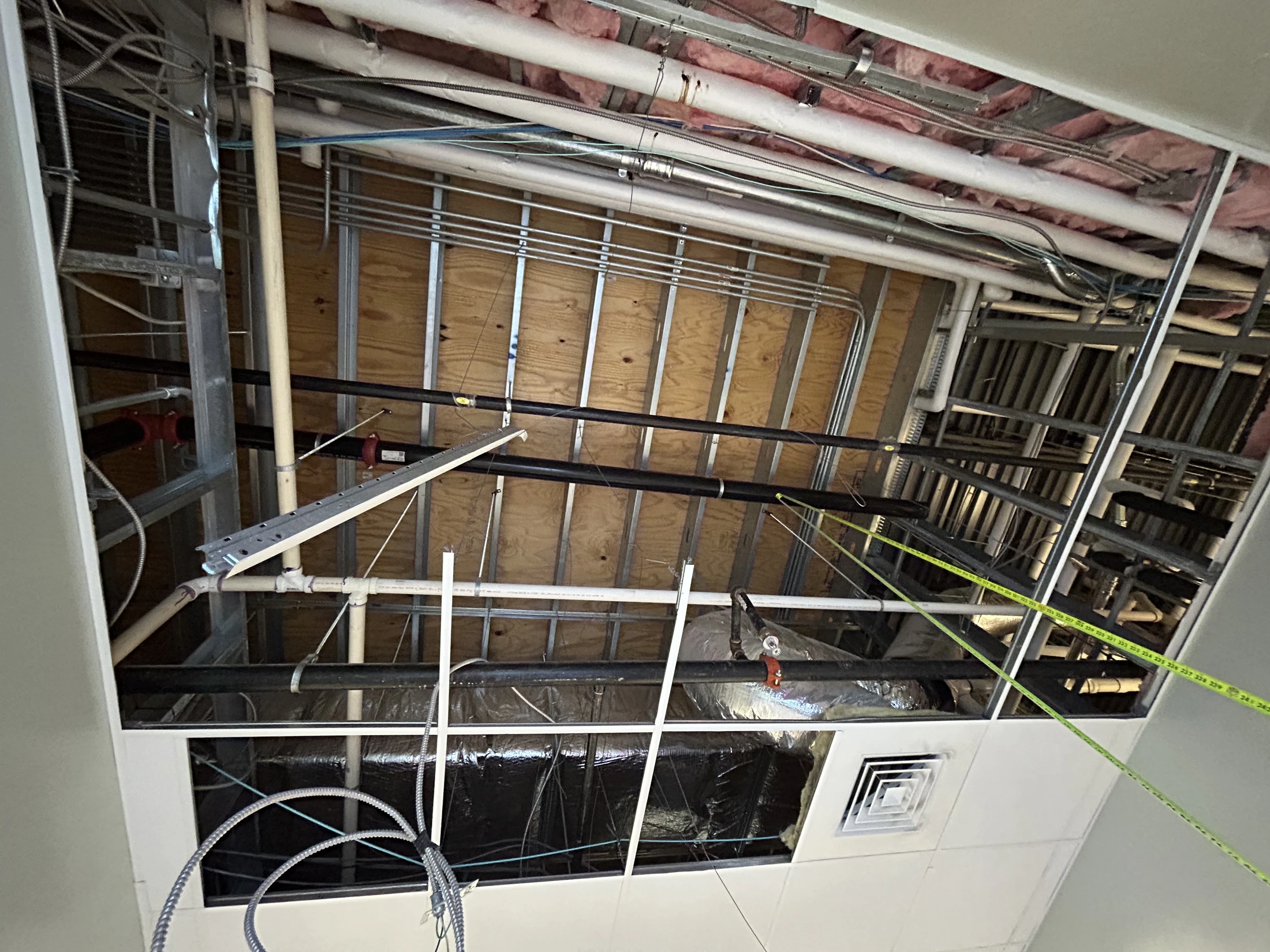A view looking up into an open drop ceiling, showing a complex network of pipes, electrical conduit, insulation, and metal framing during construction.