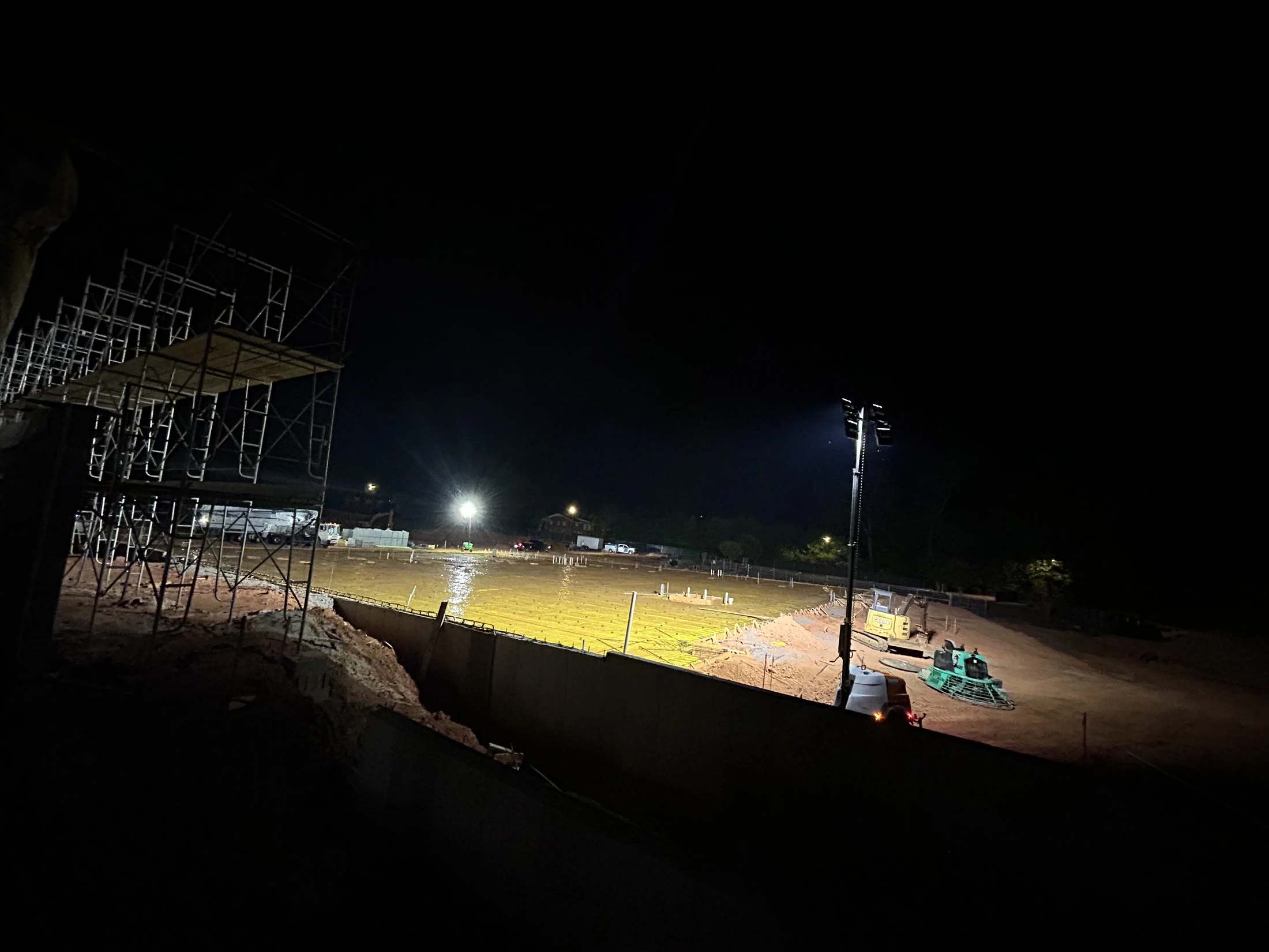 A construction site at night, with bright floodlights illuminating a large, freshly poured wet concrete slab next to scaffolding and heavy equipment.