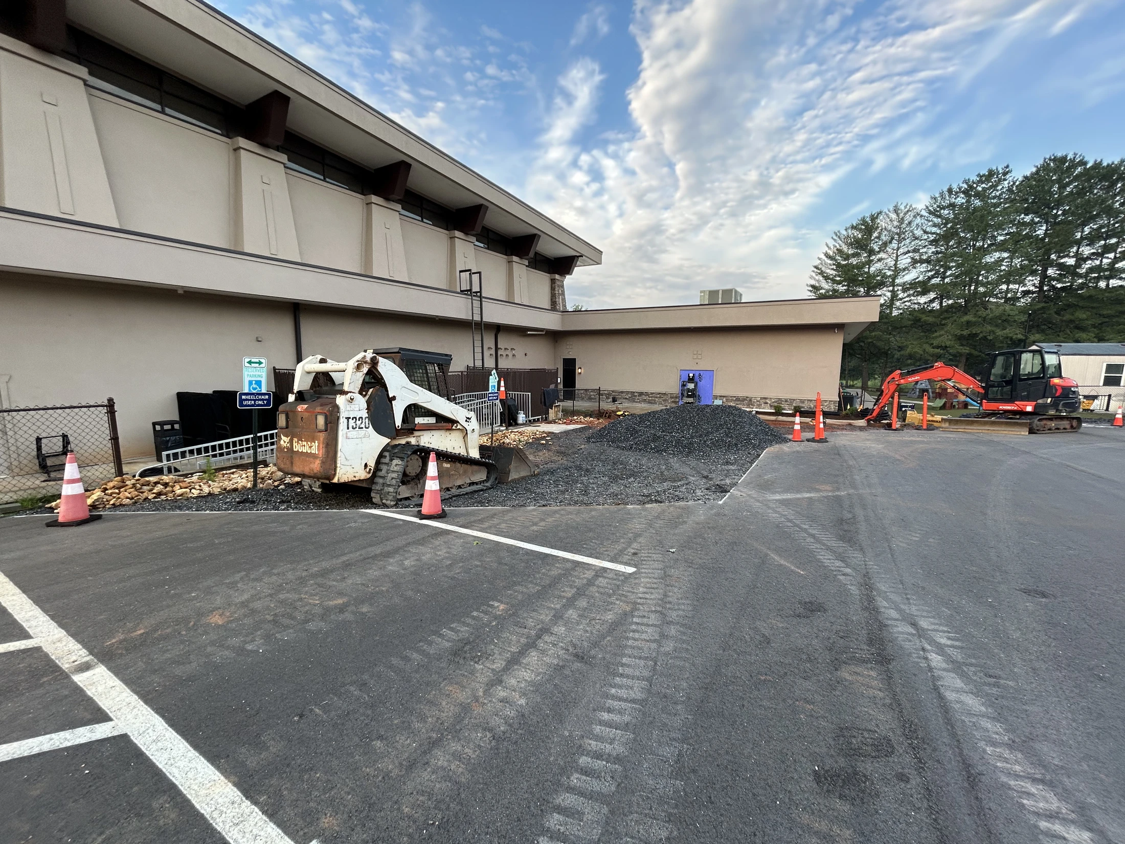 A white Bobcat loader and an orange excavator sit in a parking lot next to a building, where construction and landscaping work is underway.