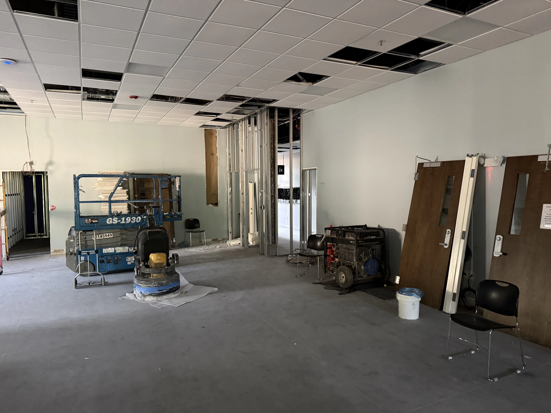 An unfinished commercial interior with a concrete floor, exposed wall studs, and an open drop ceiling. A scissor lift and a generator sit in the room.