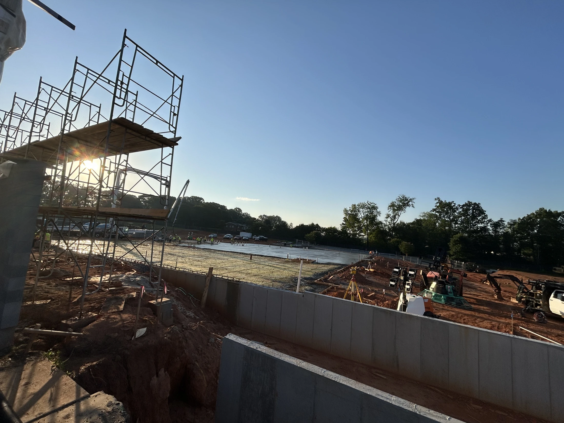 A wide-angle view of a construction site at sunrise. Scaffolding is on the left, with the sun flaring through it, overlooking a new concrete foundation.