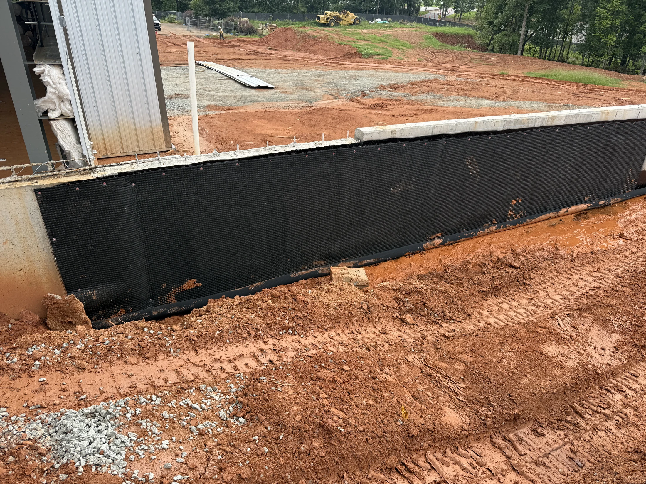A construction site with a concrete foundation wall covered in black waterproofing membrane. The ground is red clay mud with tire tracks and some gravel.